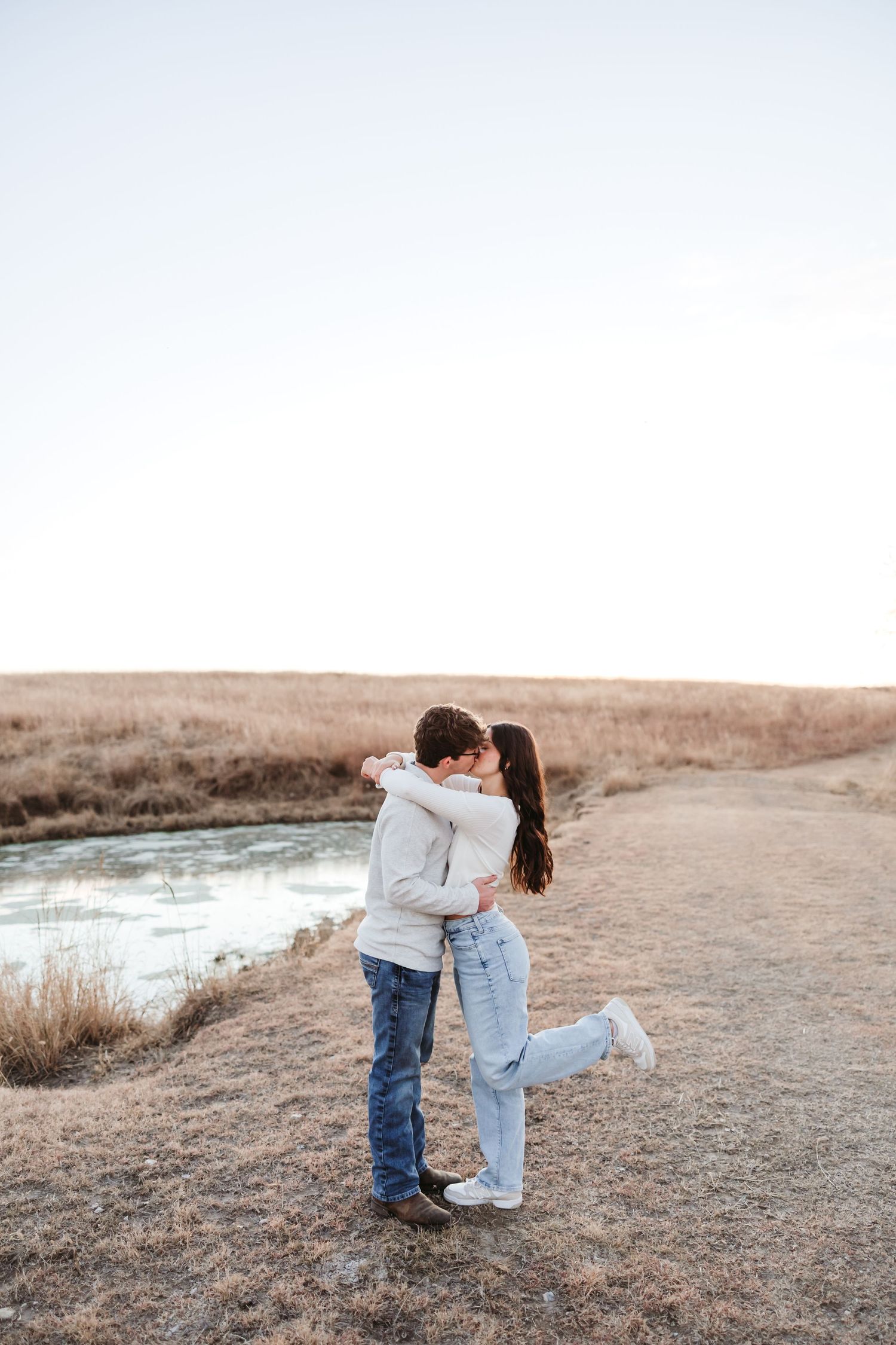 A tender embrace between two people standing on a dirt path beside a peaceful pond.