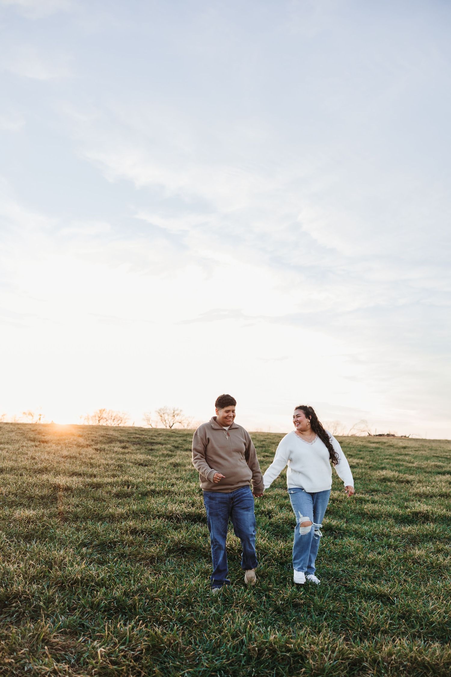Couple holding hands walking through a green field at golden hour sunset with a blue sky backdrop.