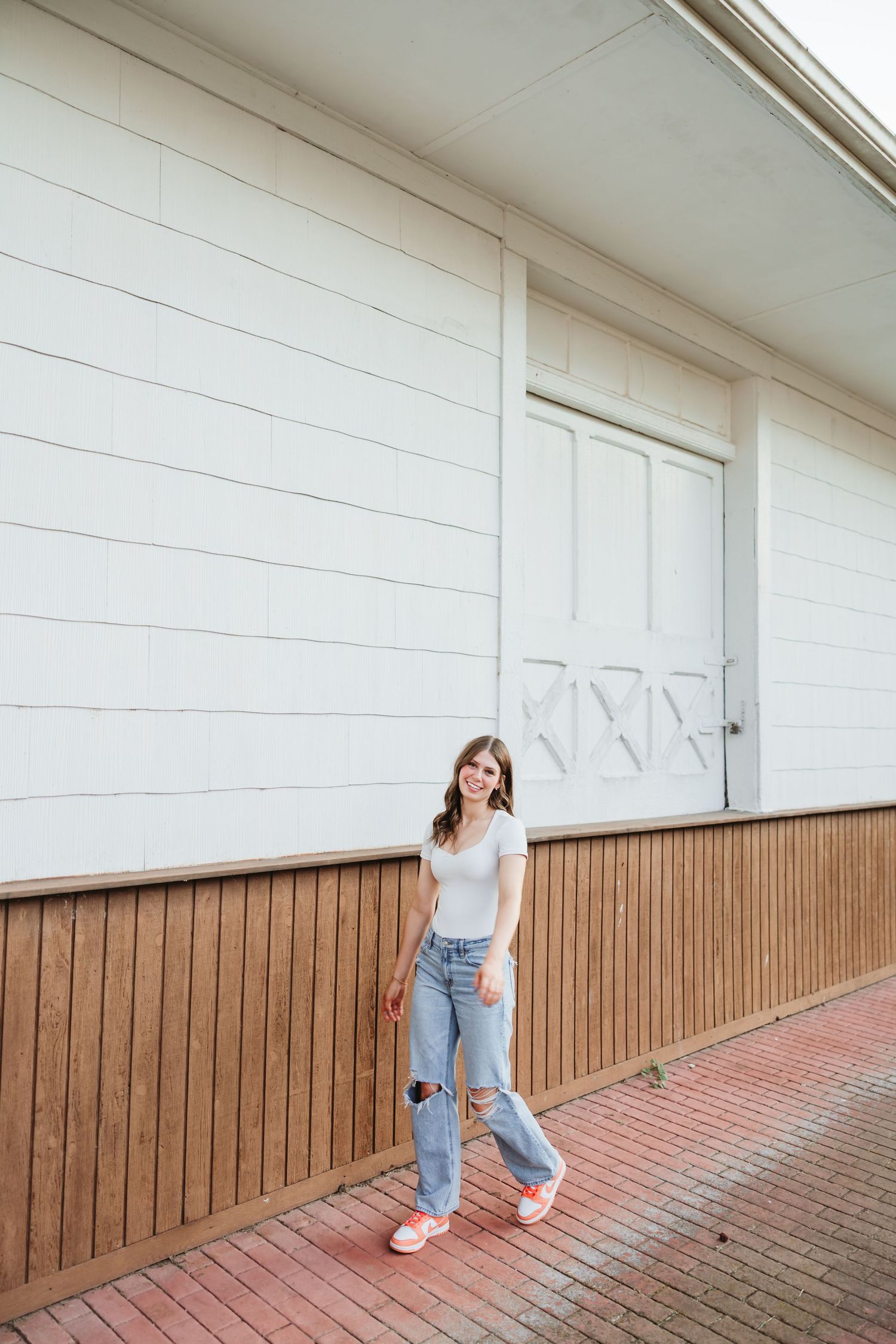 Young woman in ripped jeans and white tee poses confidently in front of a white wooden building with barn doors.