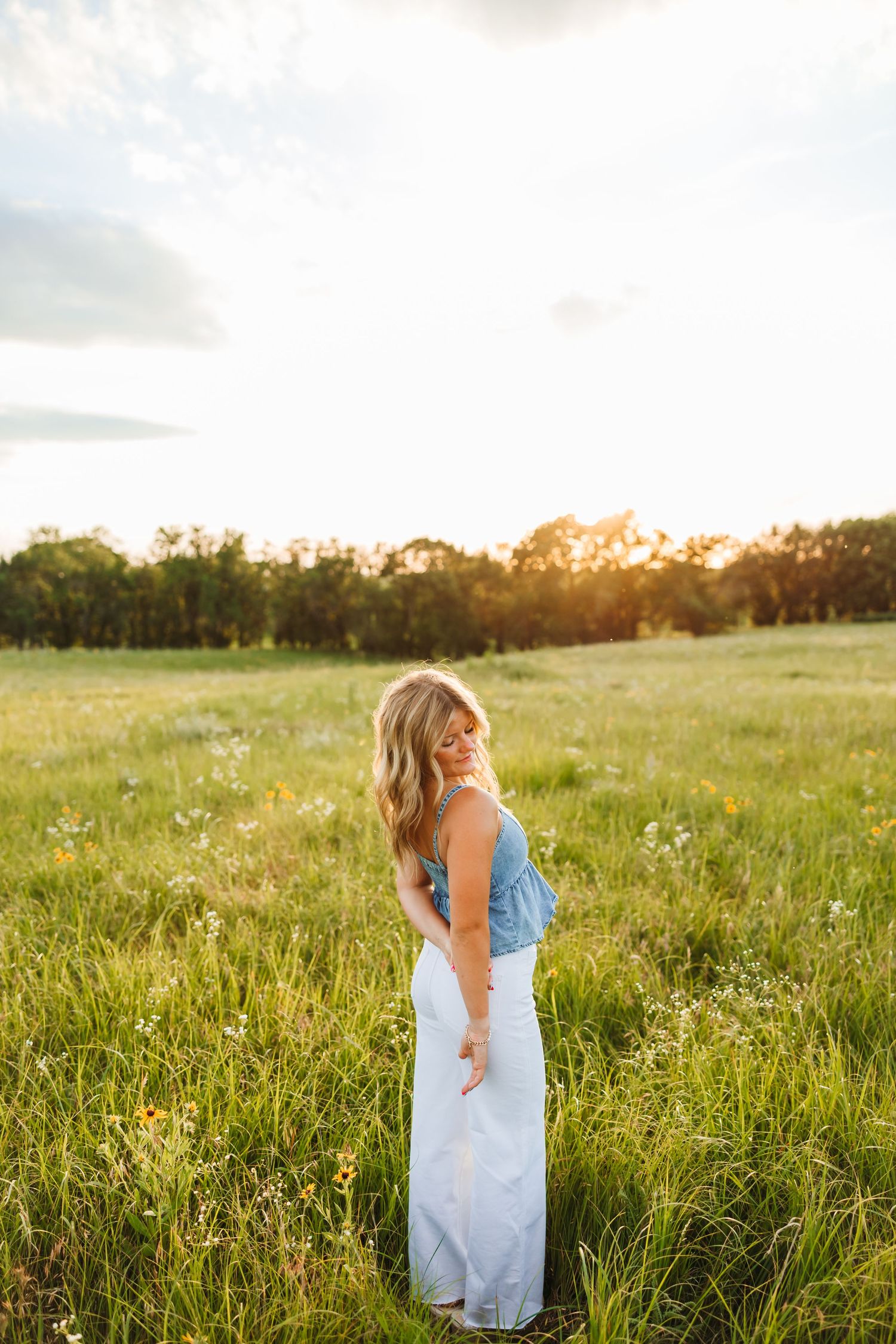 Blonde woman in blue top and white skirt standing in a sunny wildflower meadow at golden hour.
