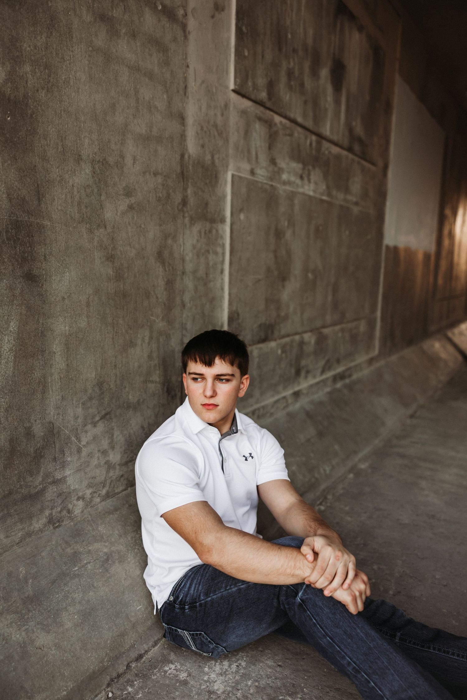 Young man in white polo shirt and jeans sitting against concrete wall outdoors.