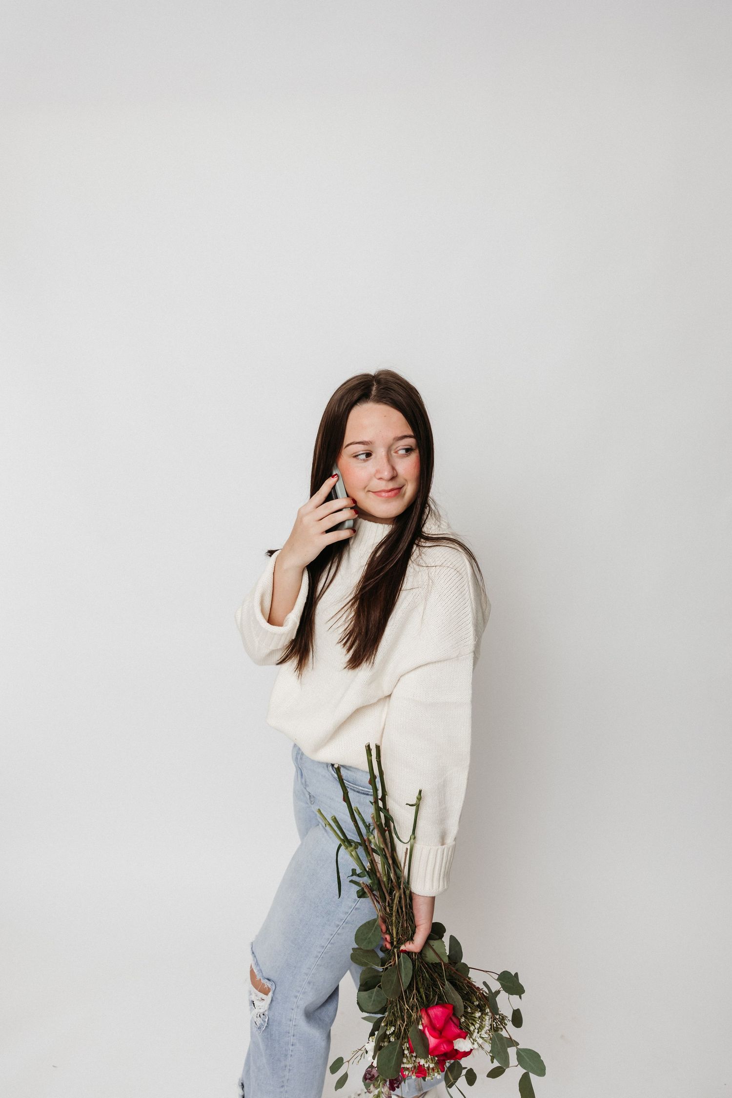 Woman in white sweater and ripped jeans poses with flowers against white backdrop.