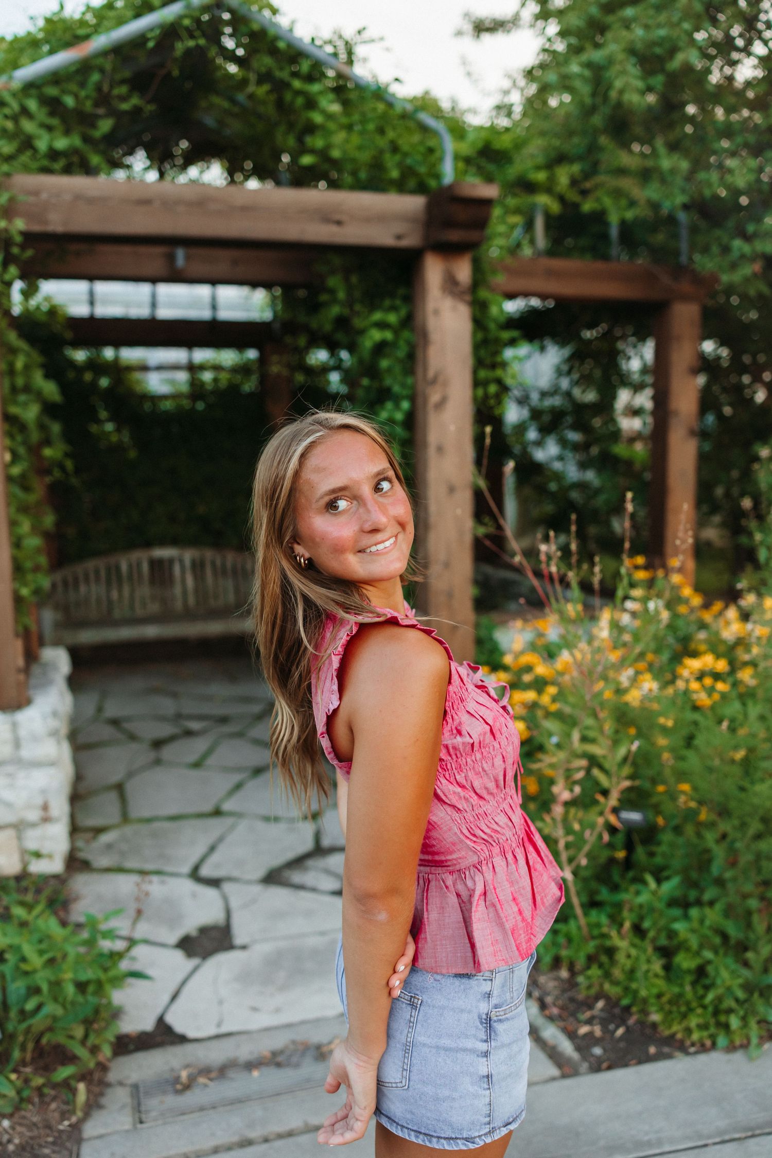 Smiling teenage girl in pink top and denim shorts poses in a garden with wooden pergola and yellow flowers.