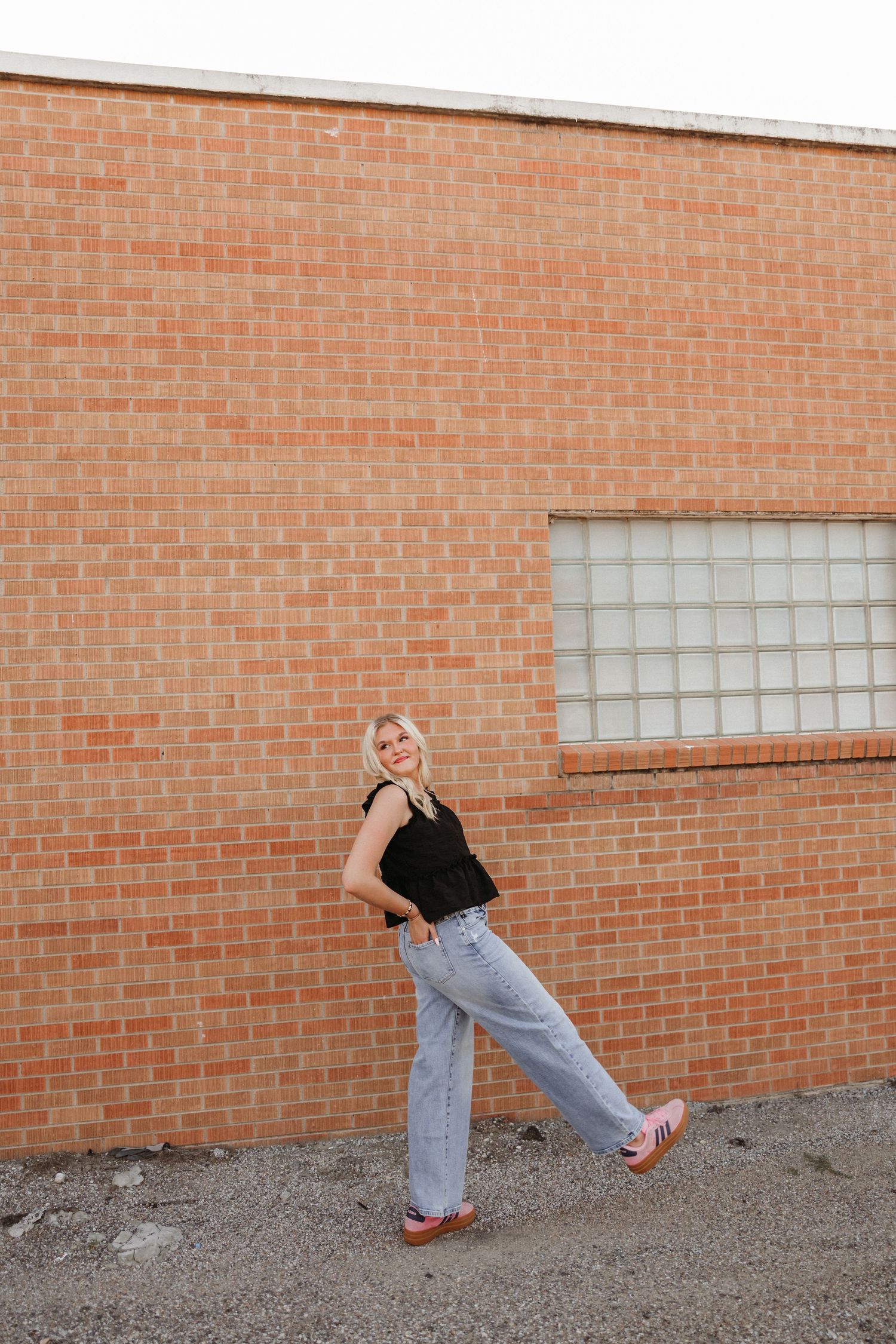 Blonde woman in black tank top and wide-leg jeans posing confidently against a red brick wall outdoors.