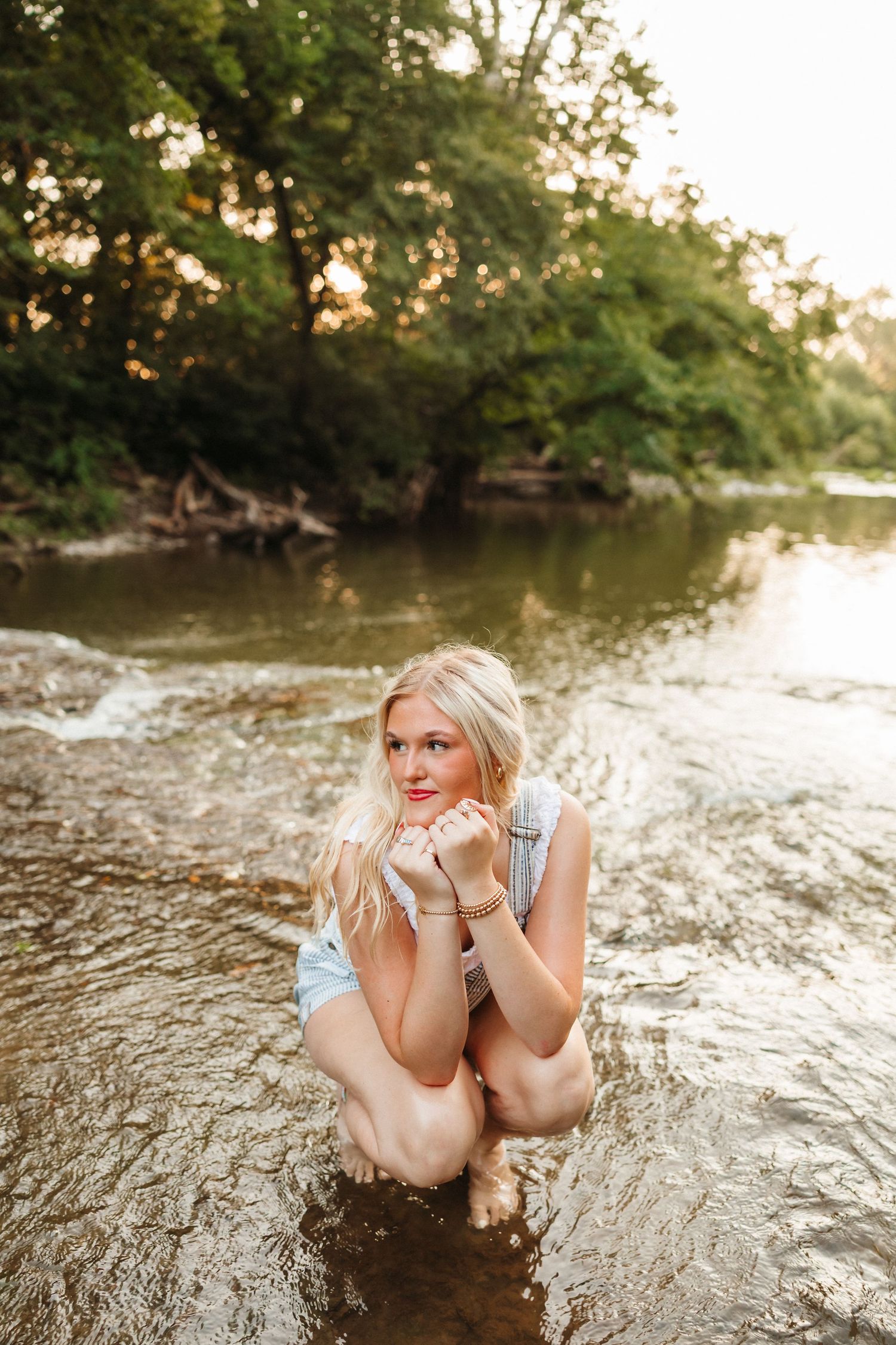 Blonde woman crouching in a shallow river surrounded by lush green trees during golden hour sunset.