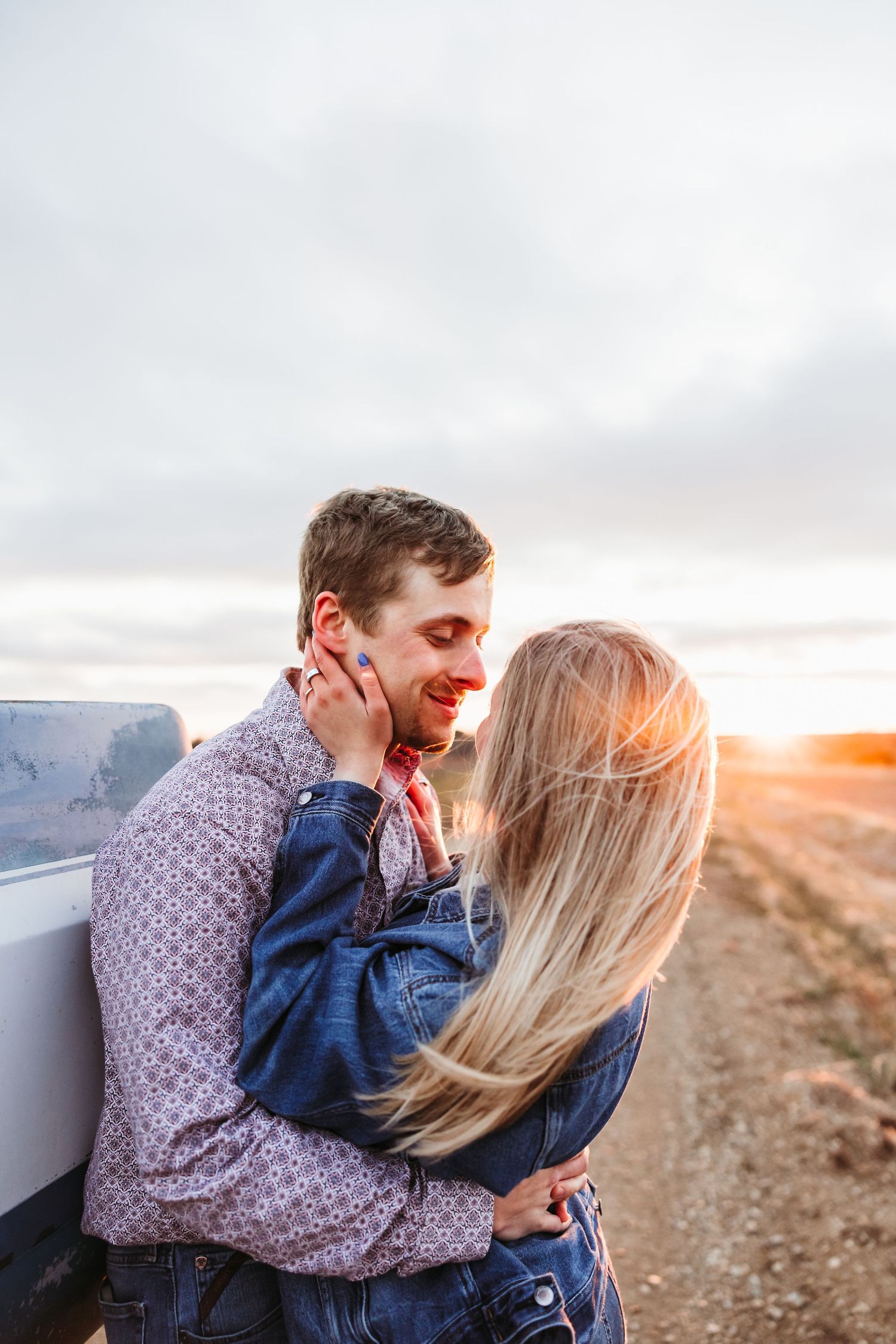 Romantic couple embracing at sunset on a rural dirt road, woman holding man's face tenderly near a vehicle.