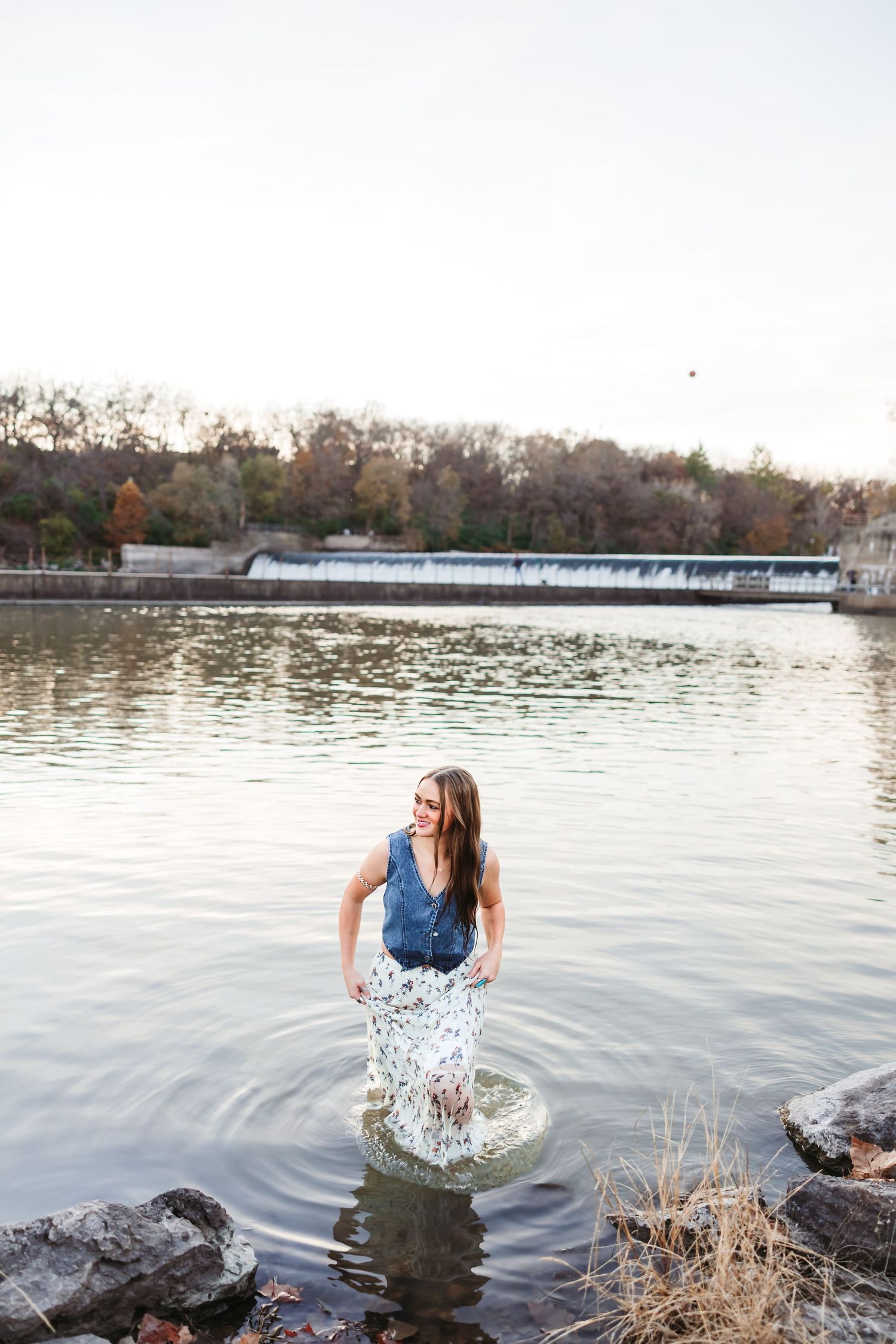 Woman standing in a river wearing a denim top and floral skirt, smiling near a dam and autumn trees.