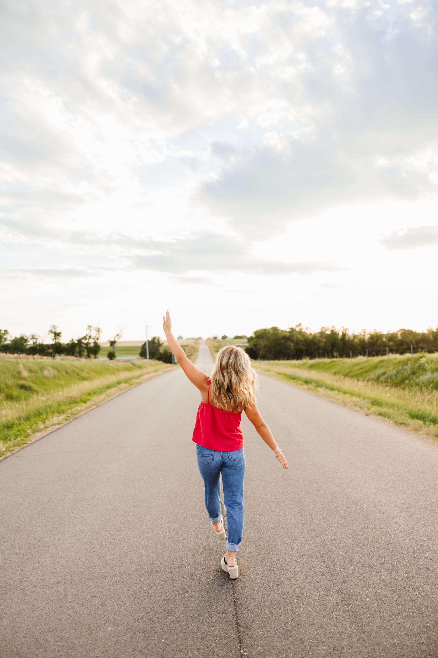 Woman in red top and jeans walking down a rural road with one arm raised toward the cloudy sky.