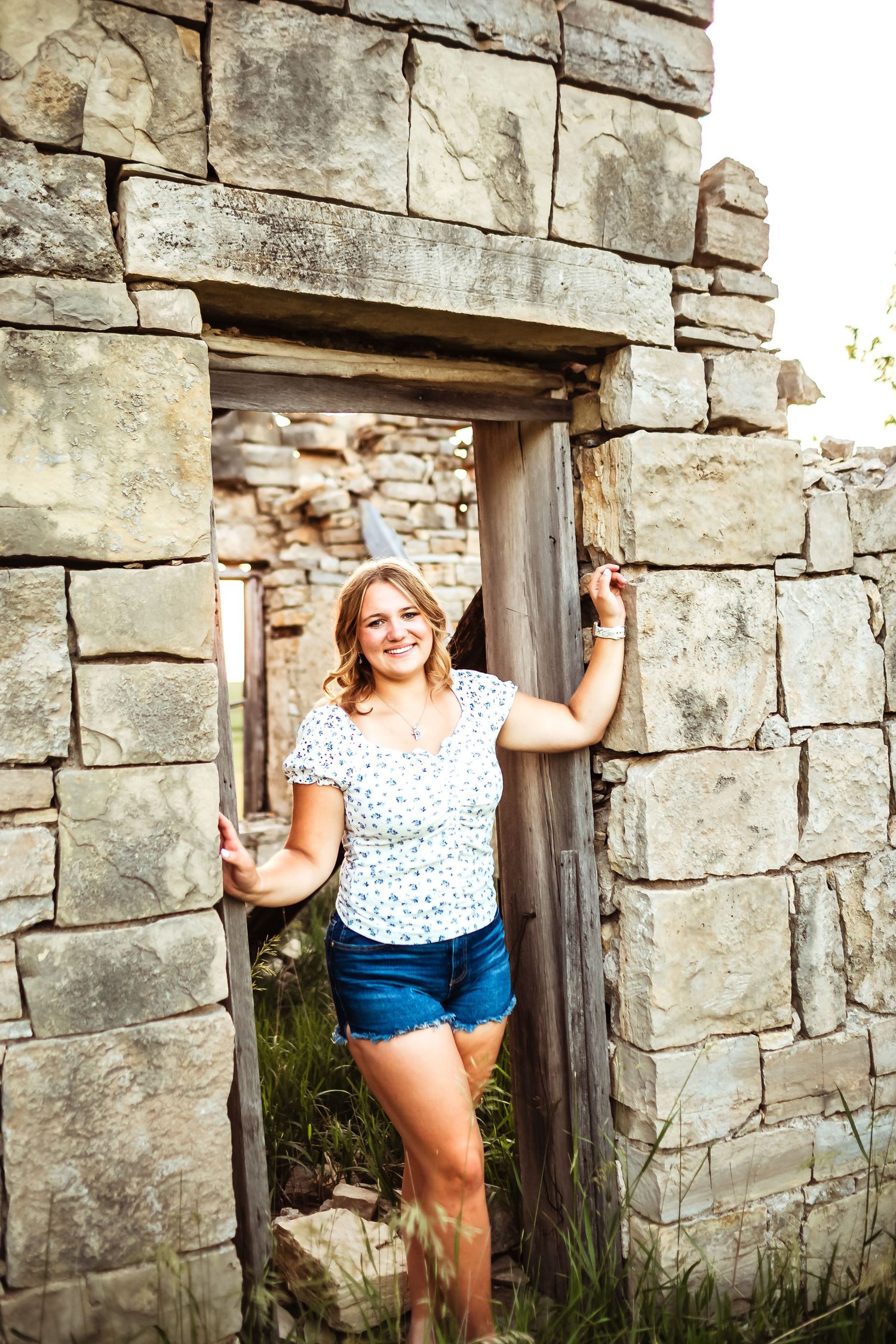 Young woman in floral top and denim shorts smiling in doorway of rustic stone ruins.