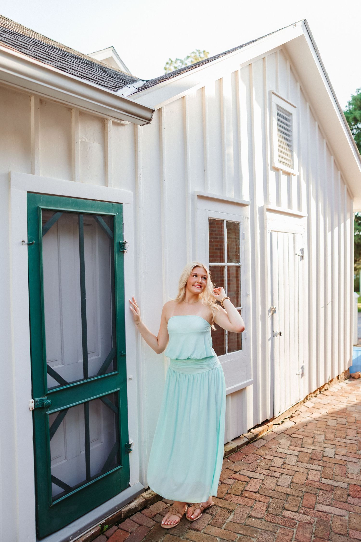 Smiling blonde woman in mint strapless maxi dress standing by green screen door of white cottage.