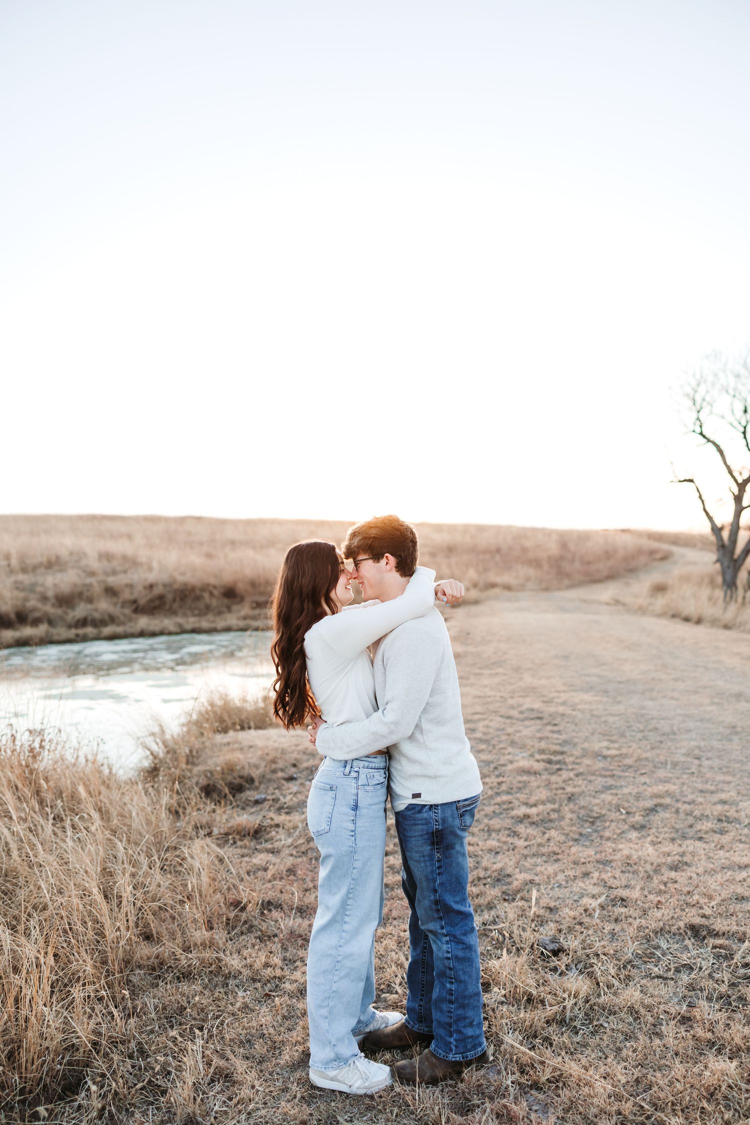 Young couple embracing and kissing in a golden field near a stream during sunset in a romantic outdoor setting.