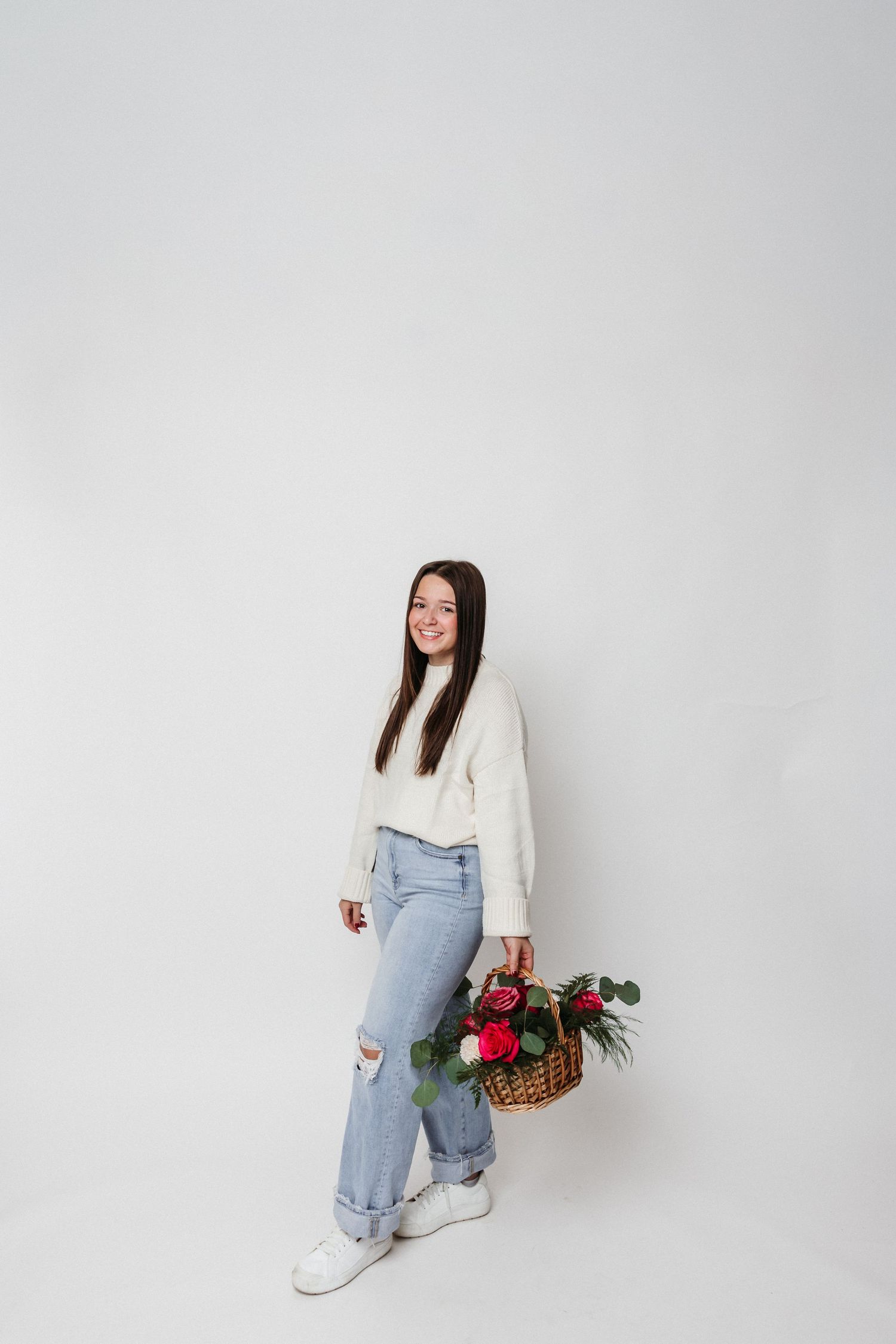 Standing pose with floral basket wearing white sweater and ripped jeans against white background.