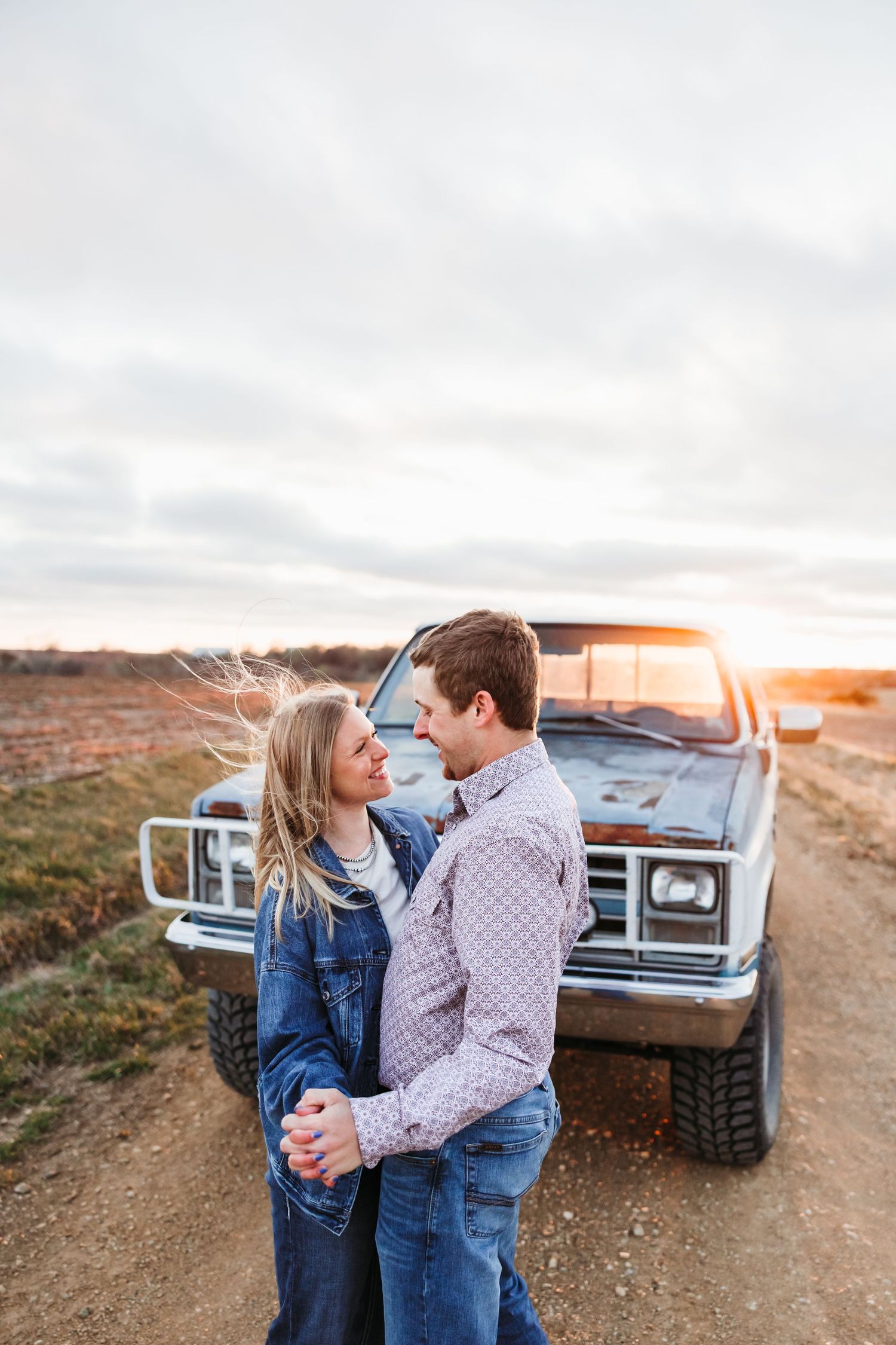 Couple embracing in front of a vintage truck on a dirt road at golden hour sunset.