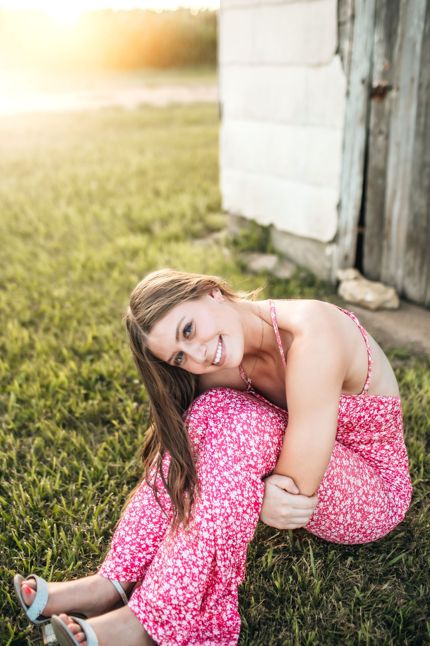 Woman in pink floral dress lying on grass near rustic building at sunset.