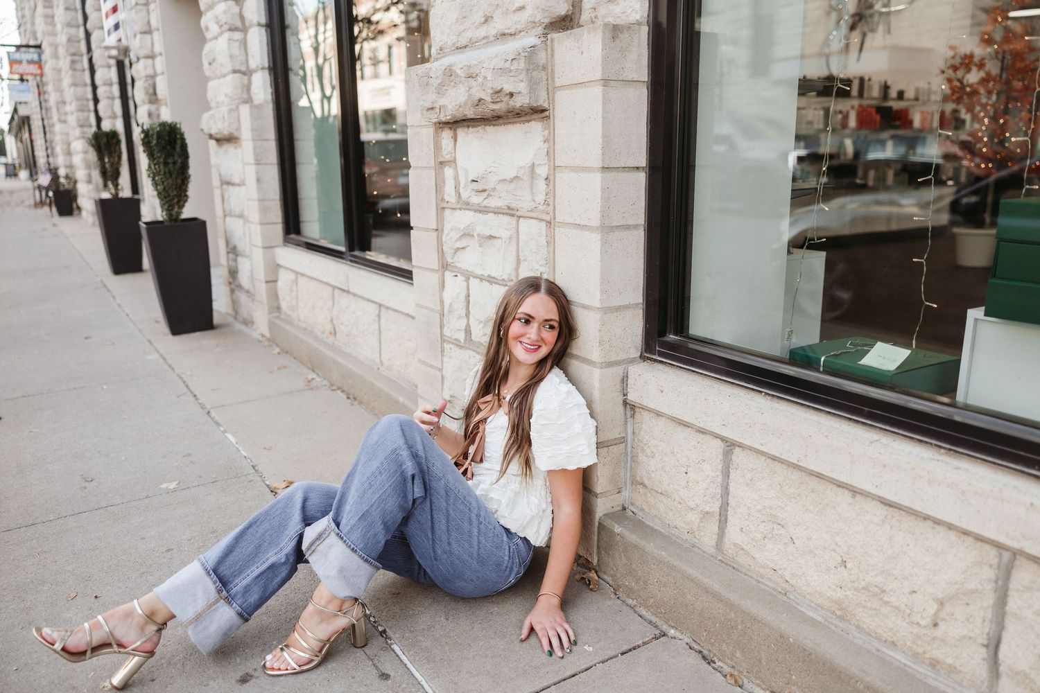 Woman in jeans and white tee relaxes against storefront window on city sidewalk.