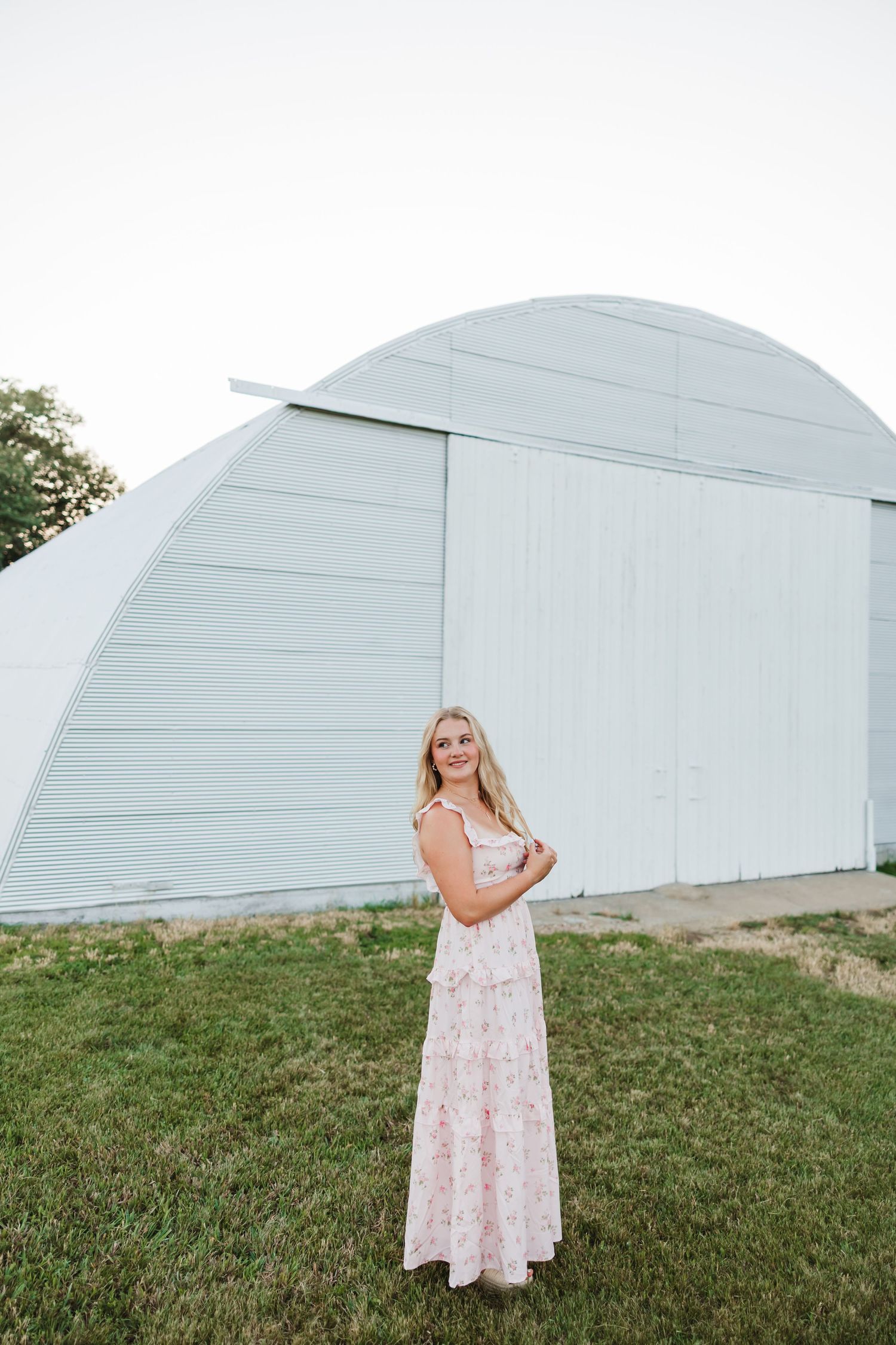 Smiling blonde woman in a pink floral maxi dress standing on green grass in front of a white quonset barn.