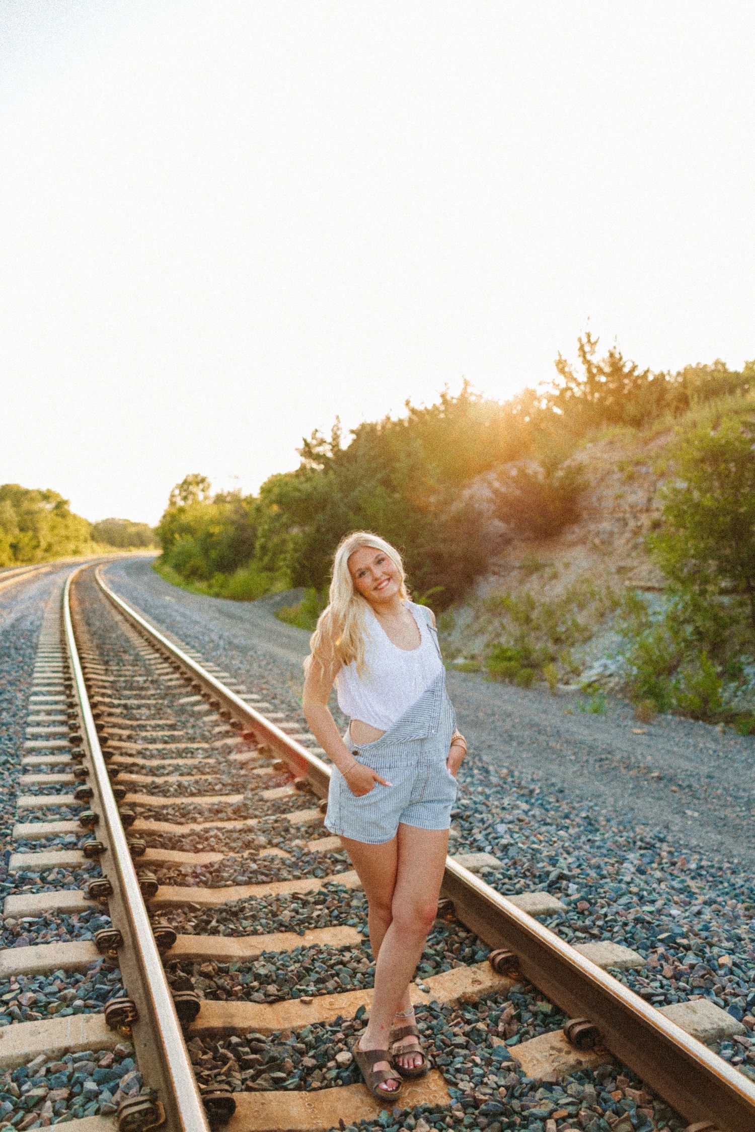Blonde teenage girl posing on railroad tracks at golden hour, wearing white top and denim overalls shorts.