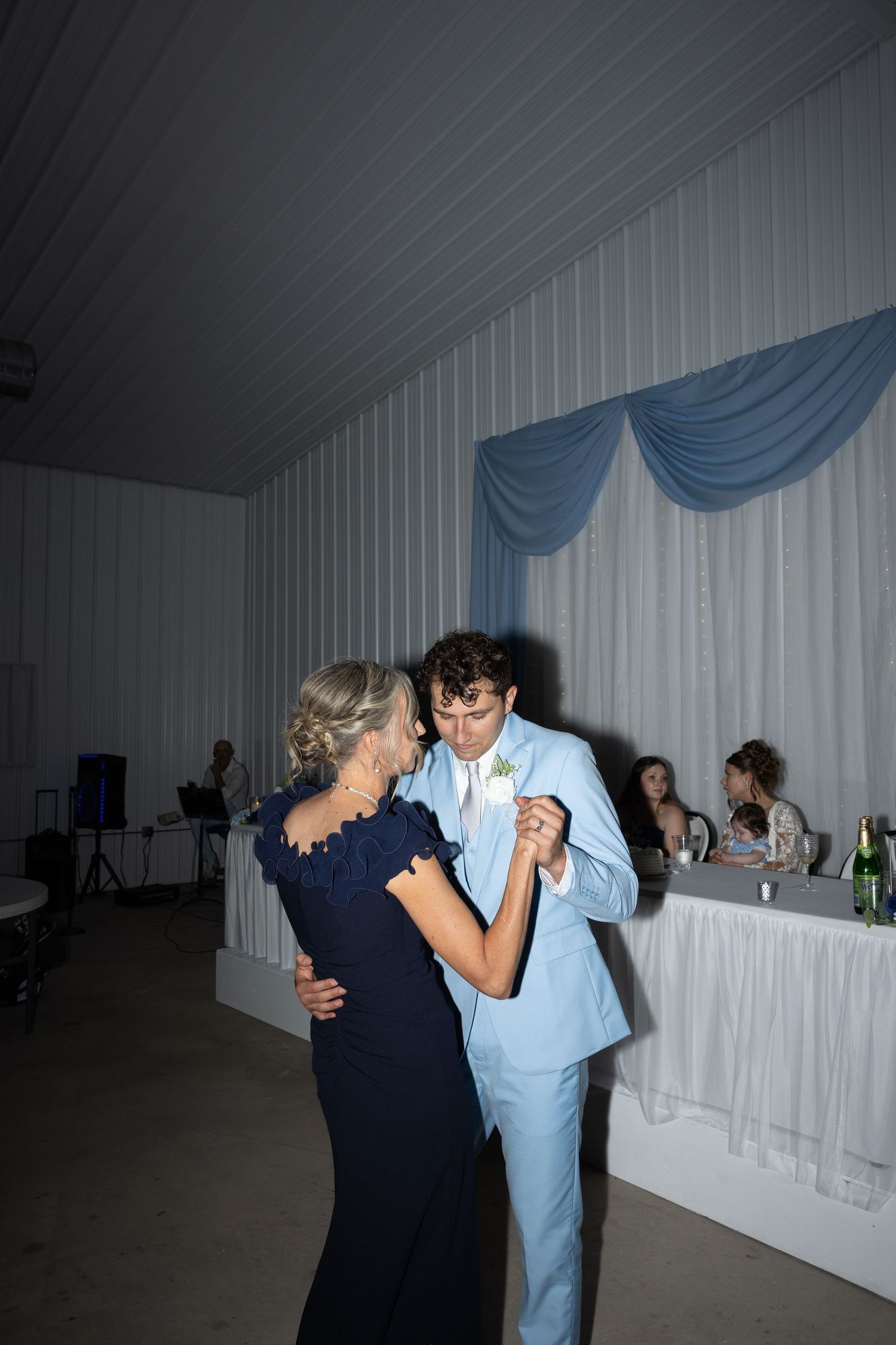 Groom in light blue suit shares mother-son dance at elegant wedding reception with blue drape decor.