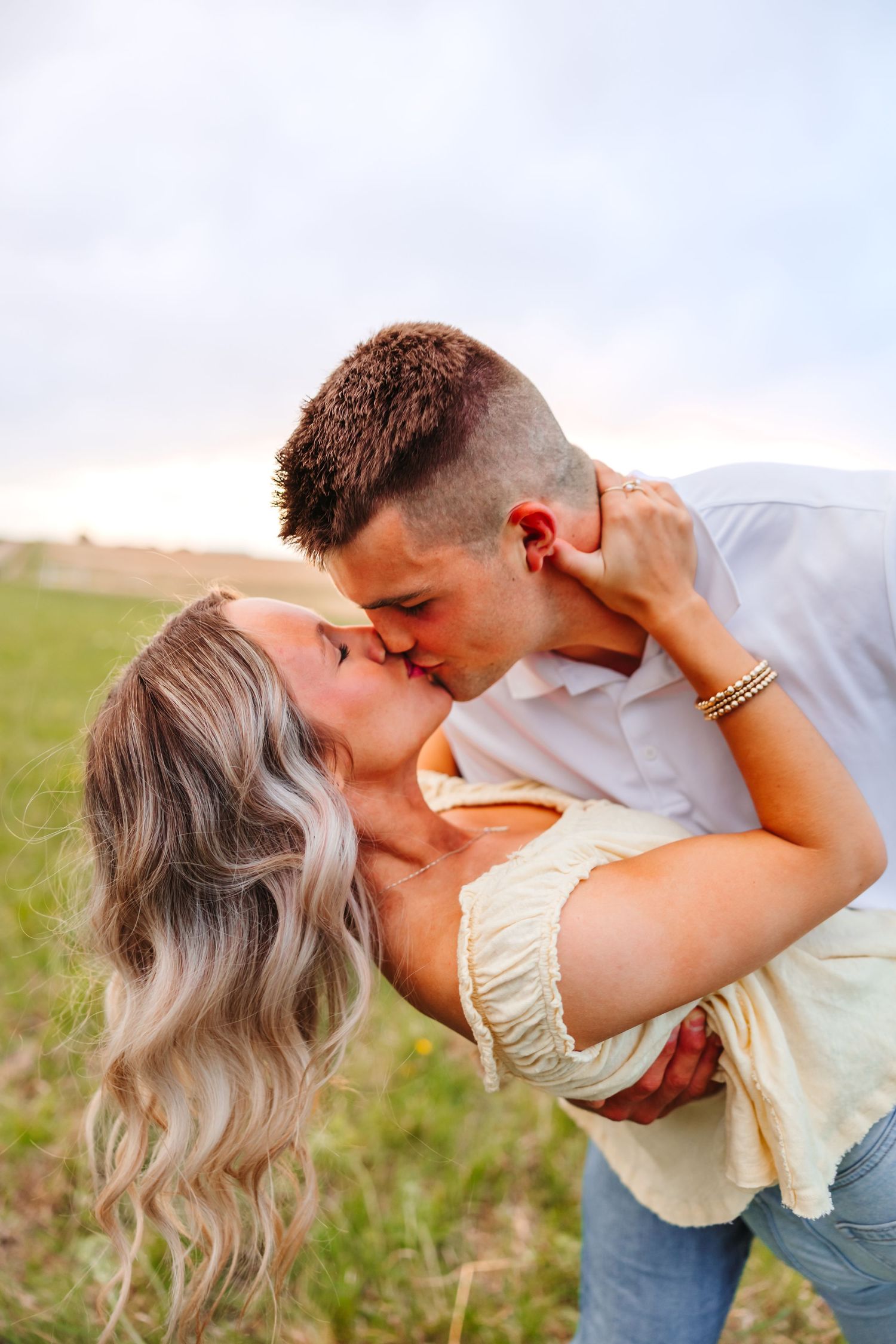A couple shares a romantic kiss in a green field, the man dipping the blonde woman as the sky glows softly behind them.