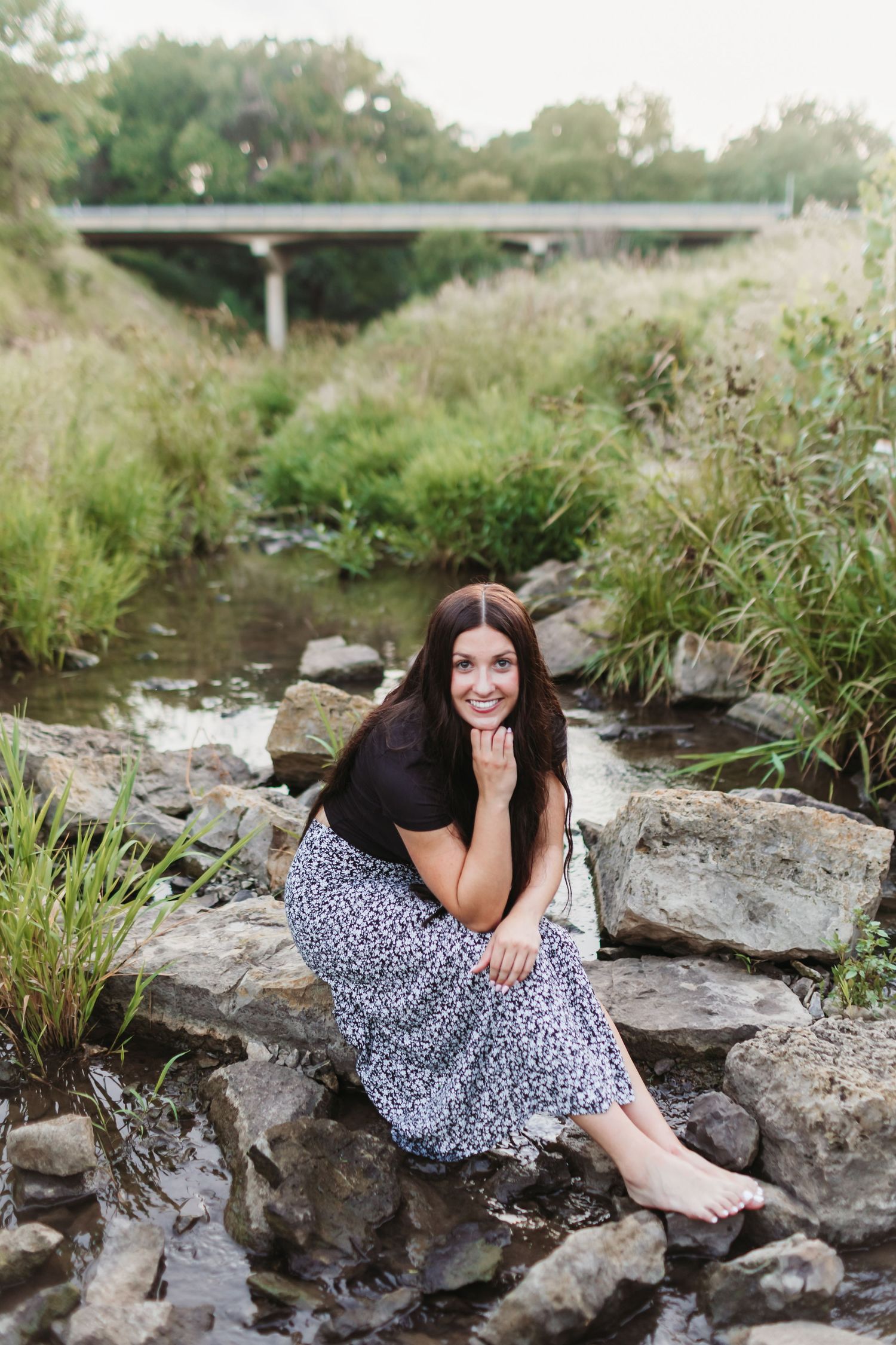 A smiling brunette woman sits barefoot on rocks by a creek, wearing a black top and floral skirt, with a bridge behind her.