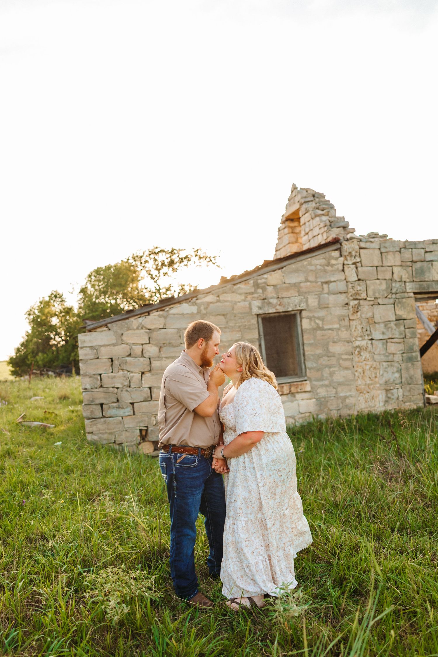 Couple sharing a romantic moment in front of a rustic stone building ruin in a green field at golden hour.