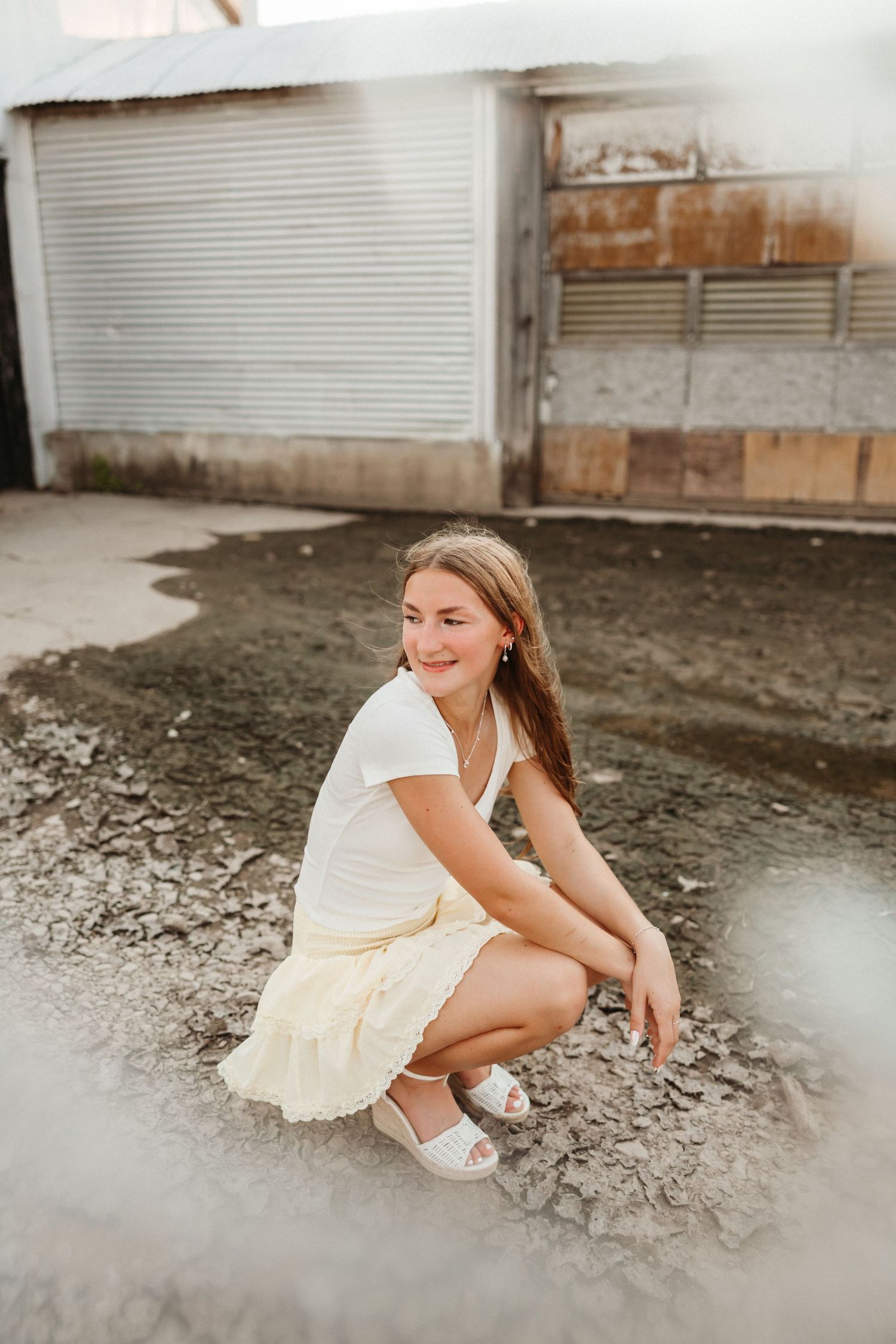 Young girl in white top and yellow skirt crouching outdoors near a building, smiling at the camera.