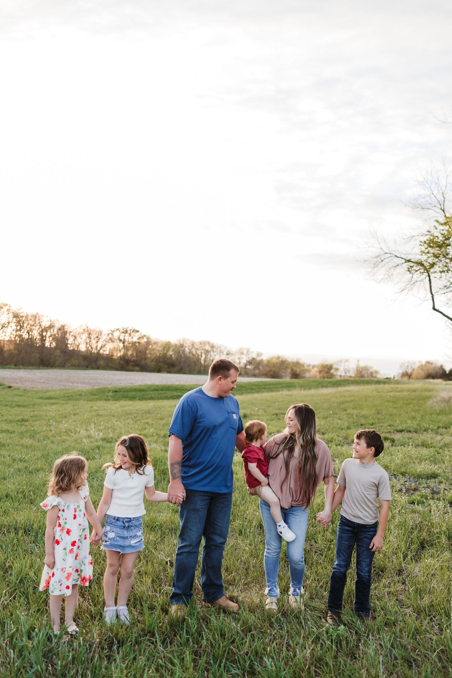 Family of six walking hand-in-hand in a sunlit green field at sunset, parents and four young children.