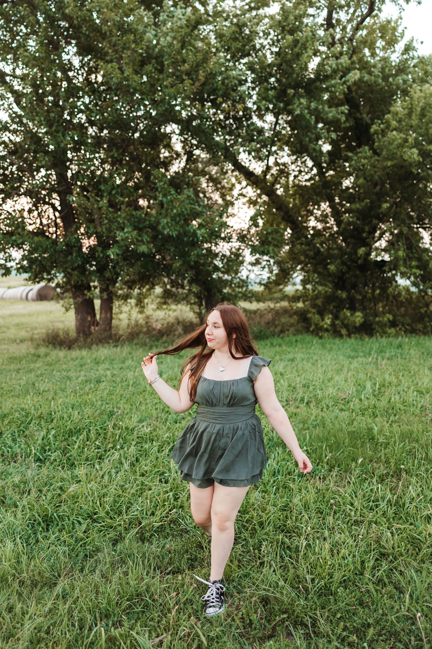 Young woman in green romper walks through lush grass field near trees at golden hour, playing with her long red hair.