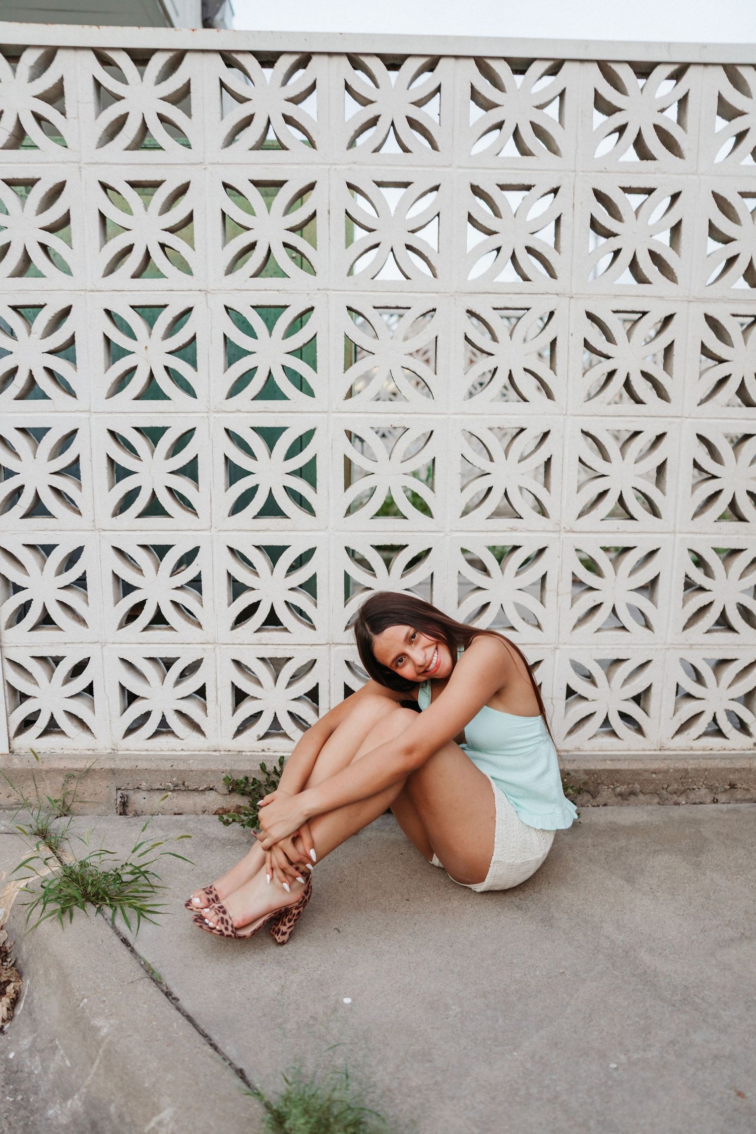 Woman in light blue top and shorts sits smiling against decorative white lattice wall on concrete ground.