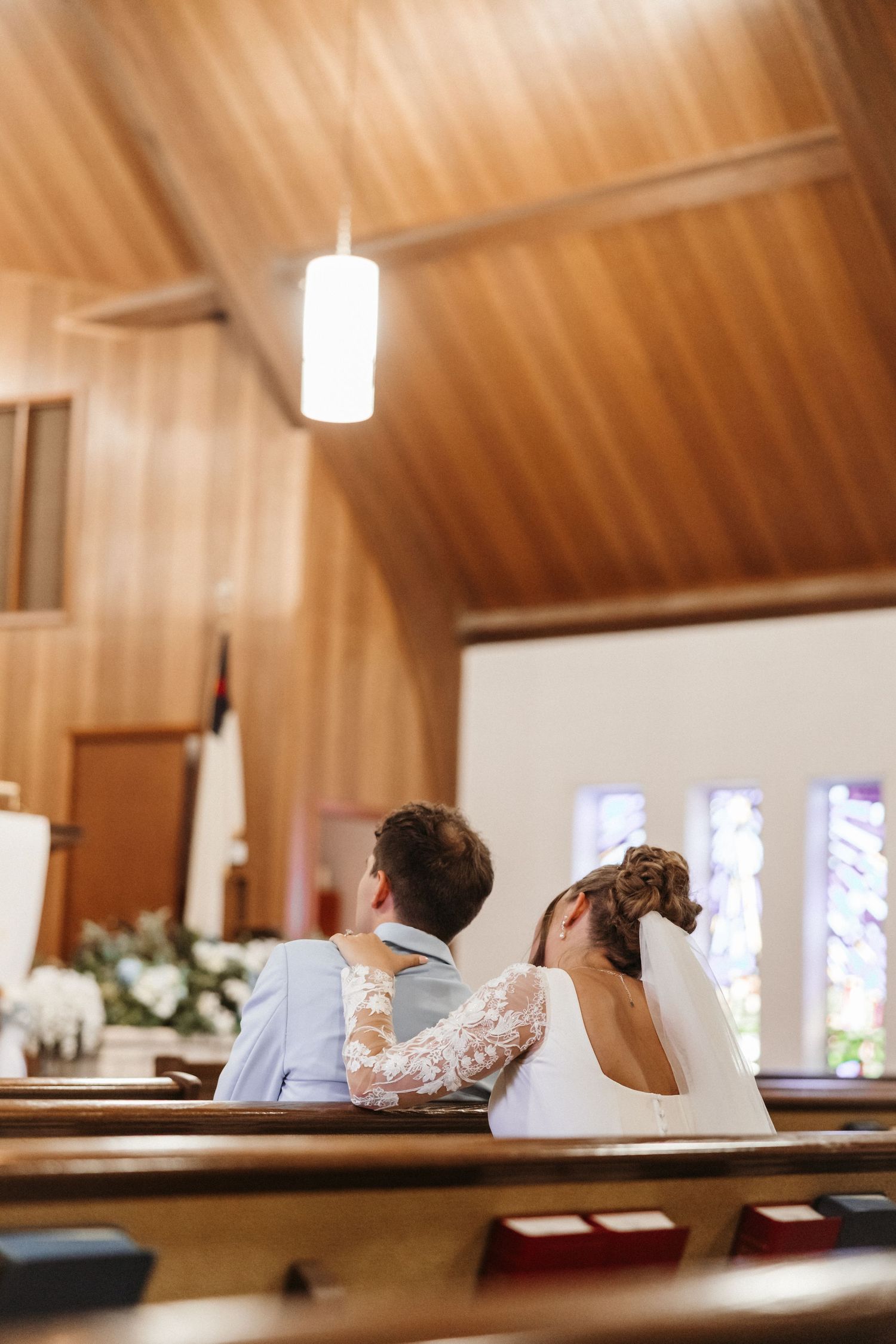 Bride and groom sitting together in wooden church pews during wedding ceremony, viewed from behind.