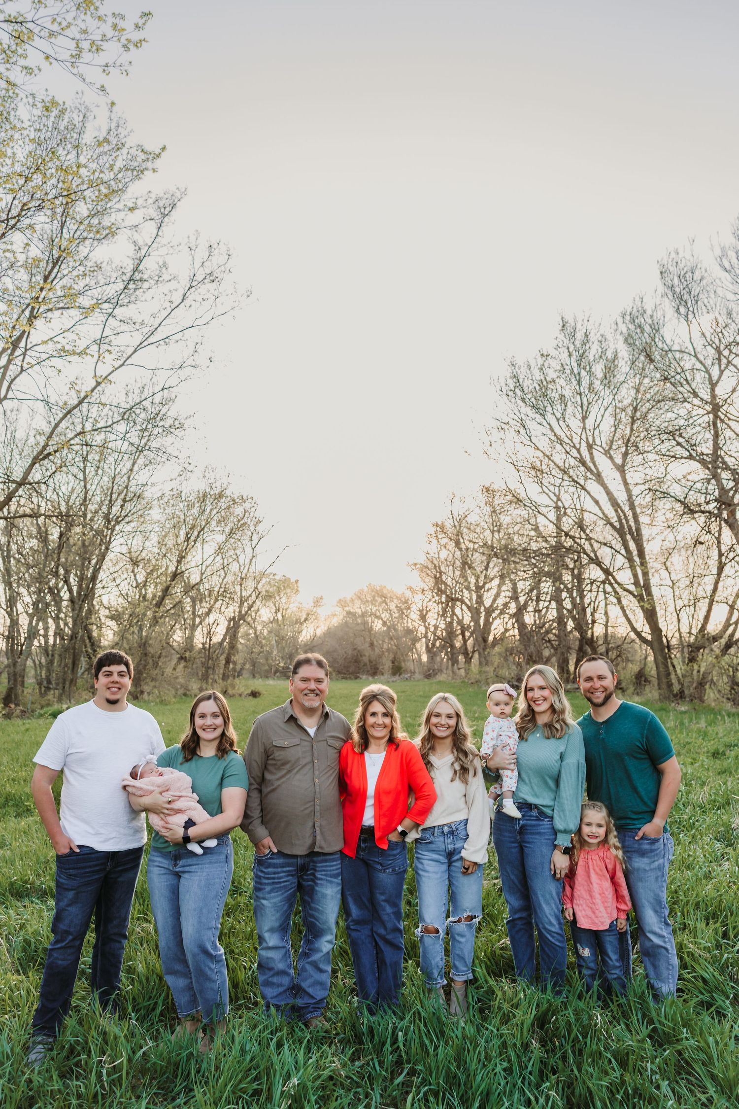 A multigenerational family of eight poses together in a lush green field surrounded by bare spring trees.