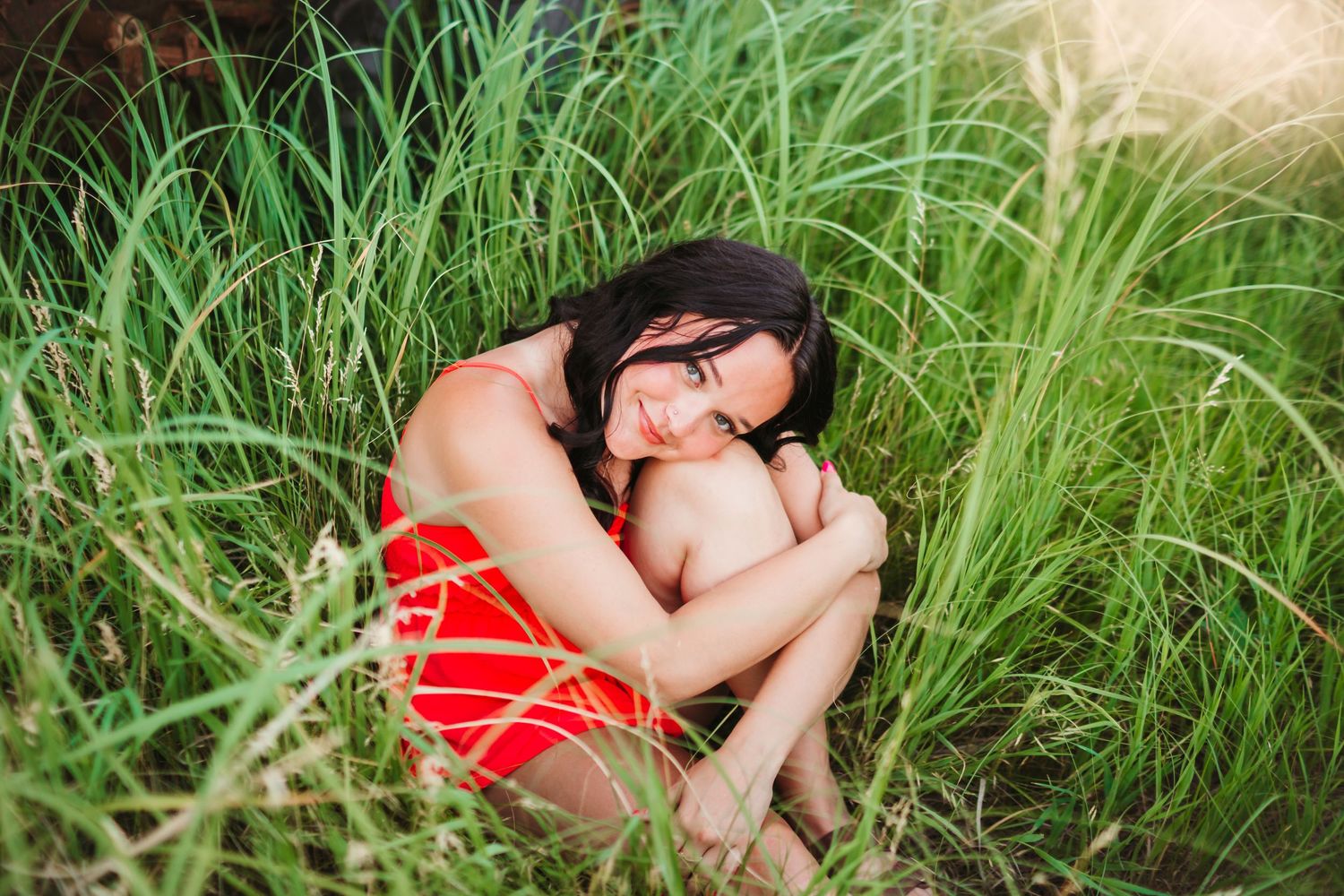 Woman in red dress curled up intimately among tall green grass.