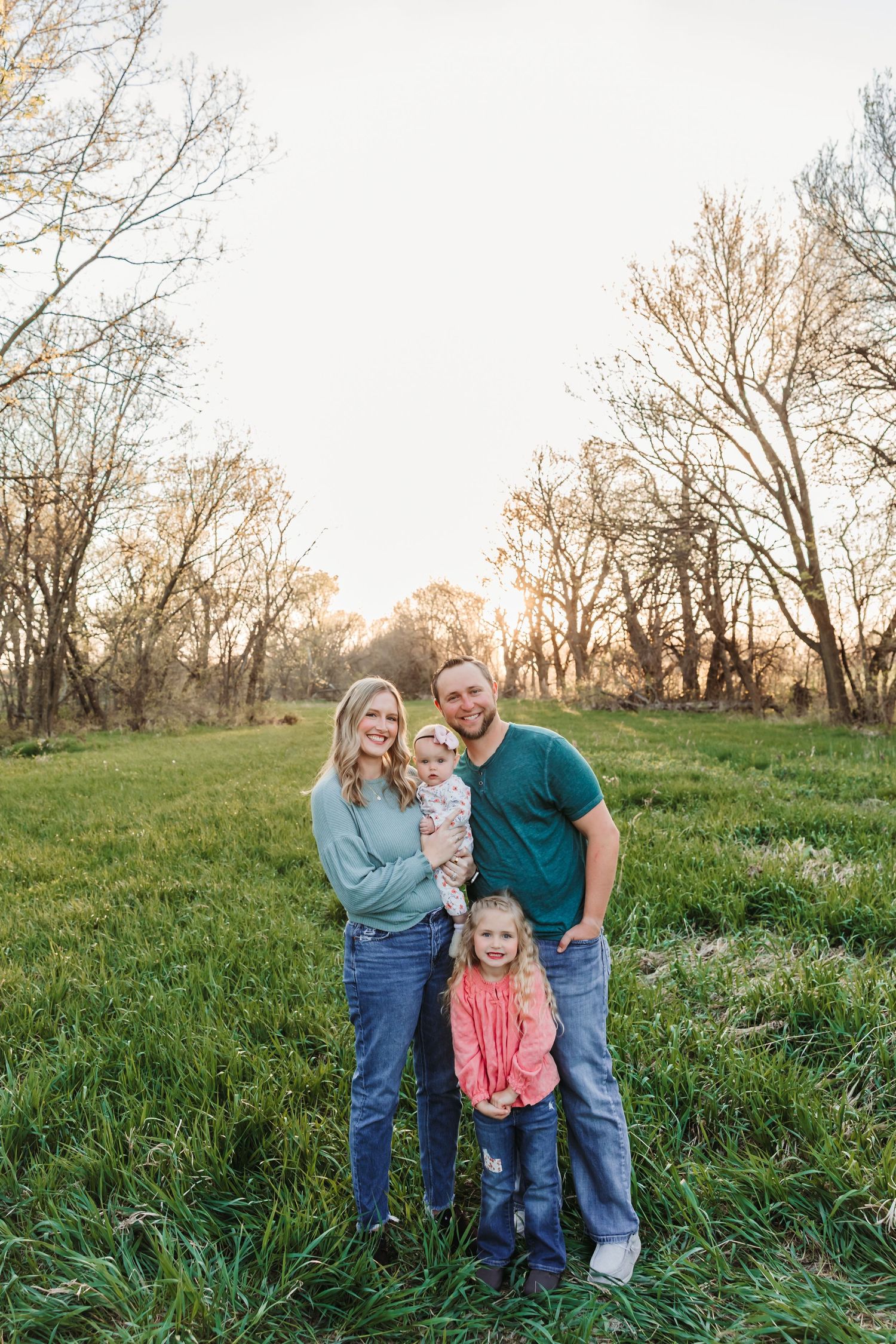 Young family of four posing in a green meadow at golden hour, surrounded by bare trees in a serene outdoor setting.