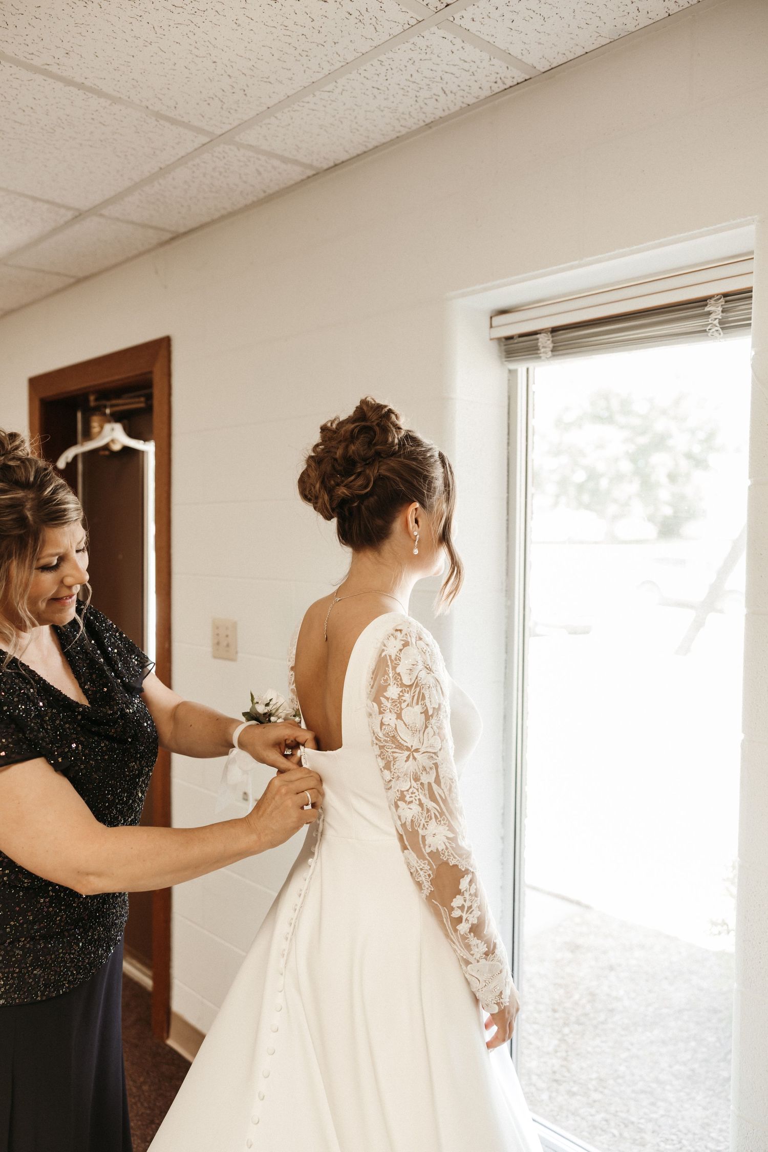 A bride in a lace long-sleeve gown gets her dress fastened by a woman in a dark dress near a bright window.