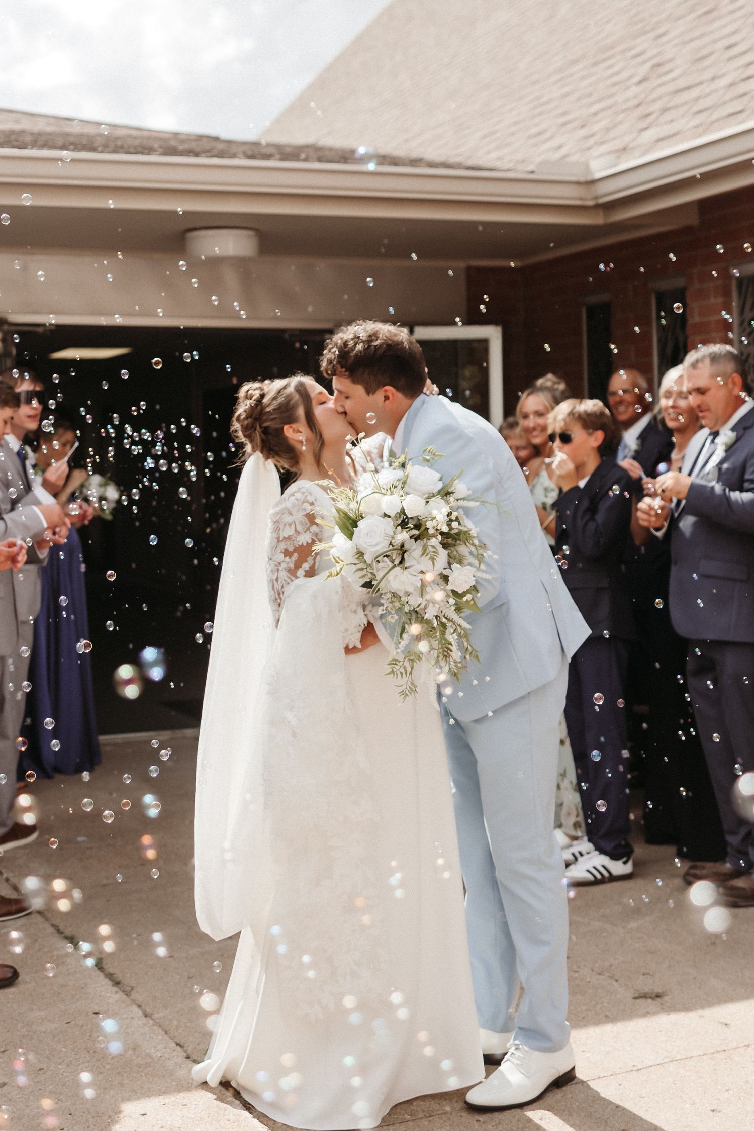 Bride and groom share a kiss surrounded by bubbles outside a church, bride holding white floral bouquet.