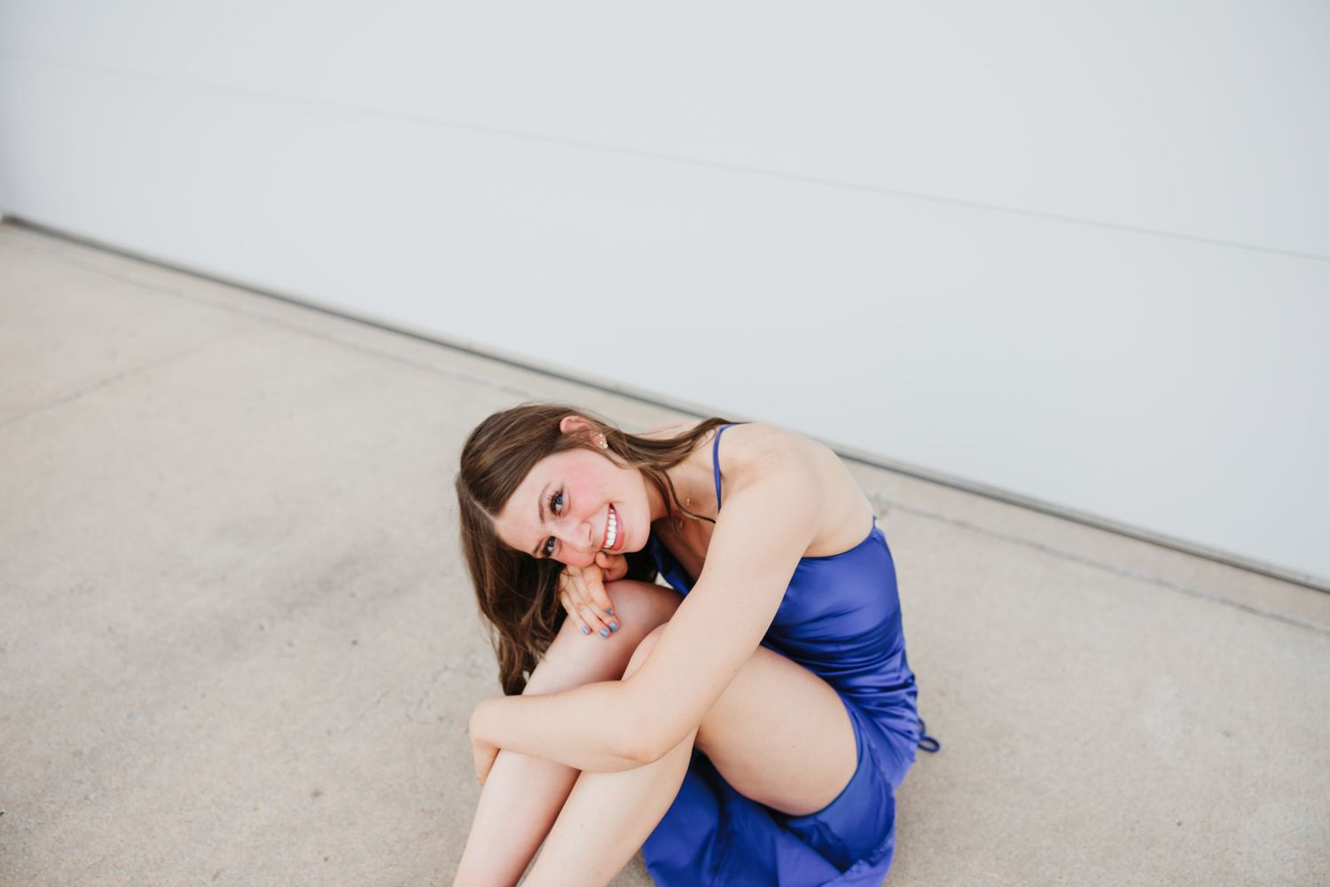 Woman in blue dress sits curled up against white wall with joyful expression.