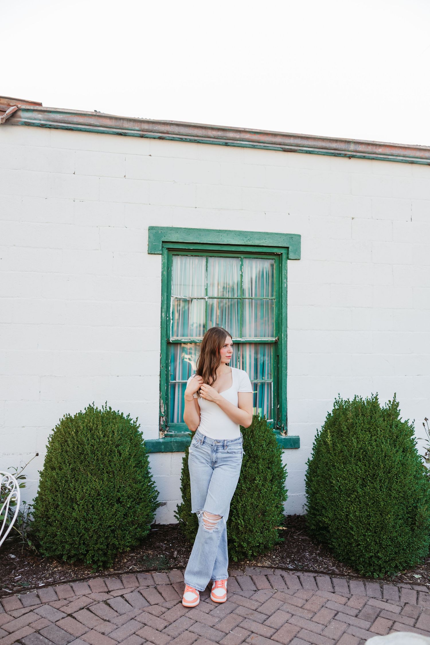 Woman posed in front of white building exterior with green-trimmed windows and decorative shrubs.
