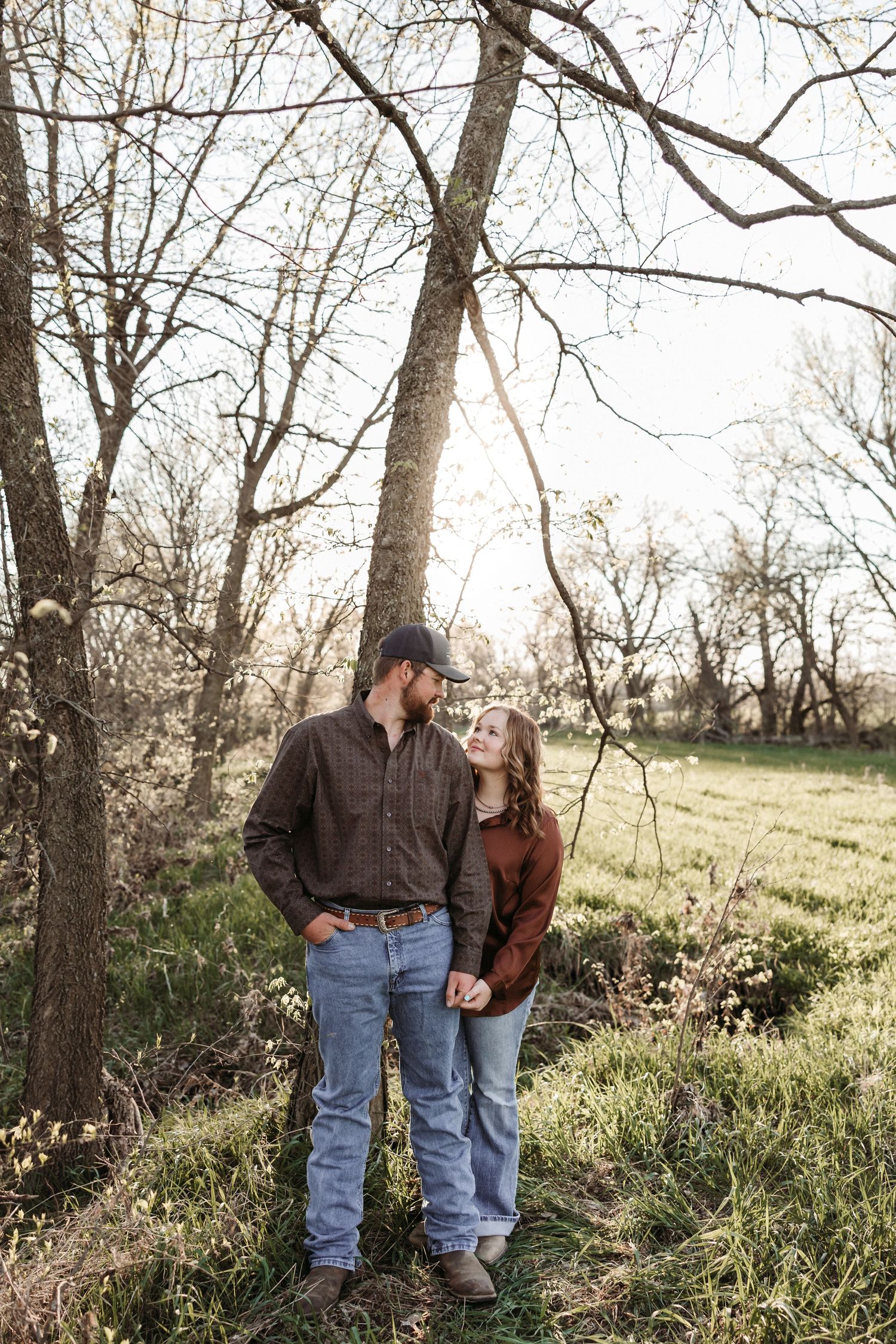 Couple holding hands and gazing at each other under bare trees in a green field during golden hour.