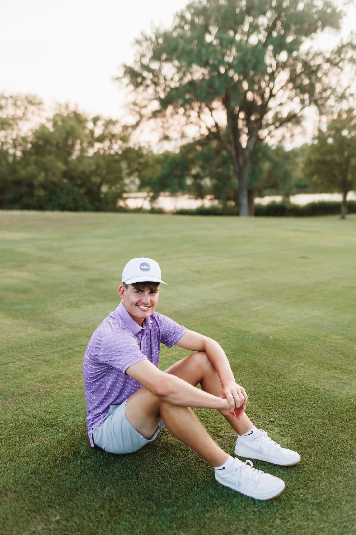 Young man in purple polo and white cap sitting on golf course green, smiling casually in summer attire.
