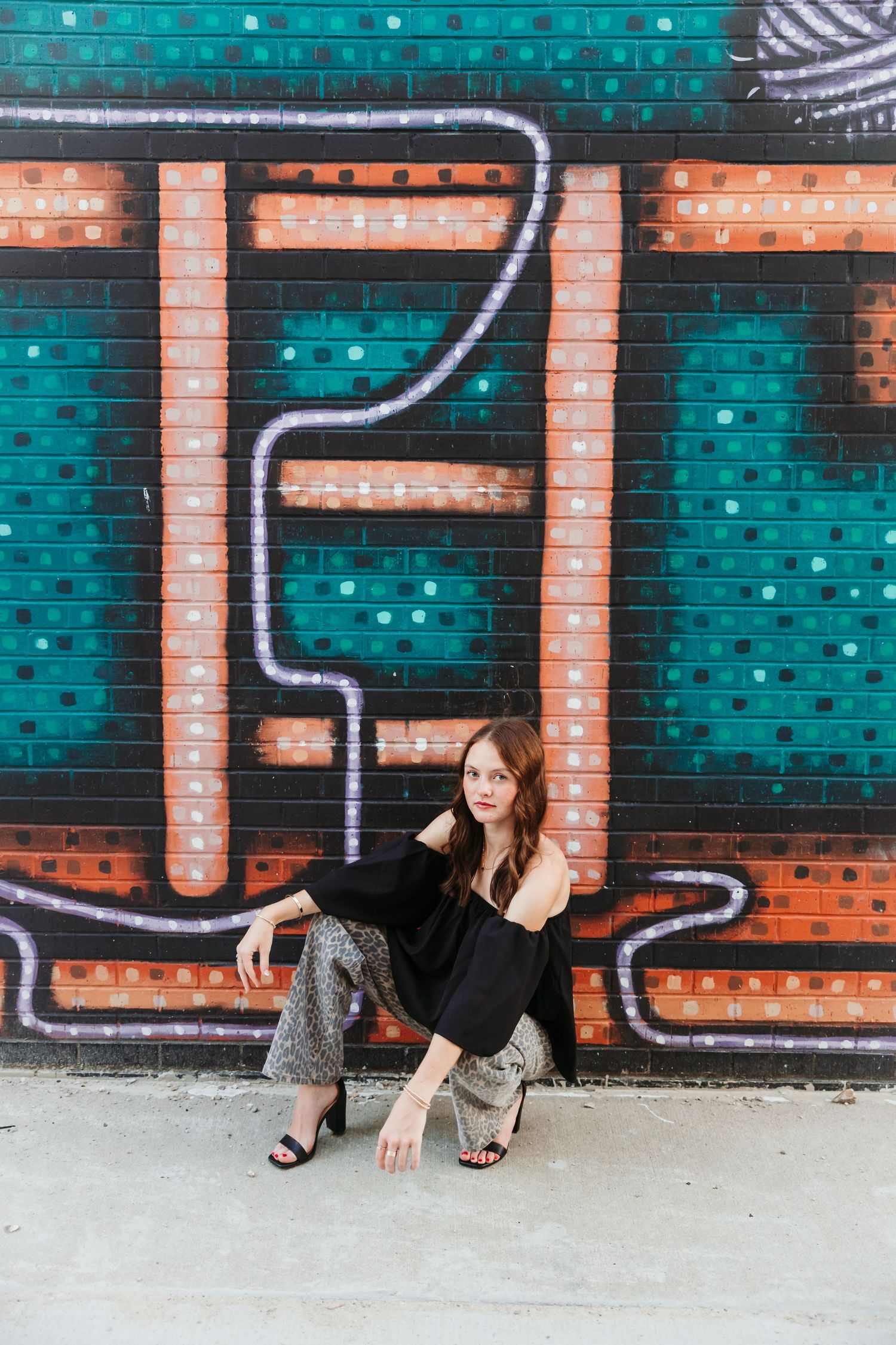 Young woman in black top and patterned pants posing against a colorful graffiti mural wall.