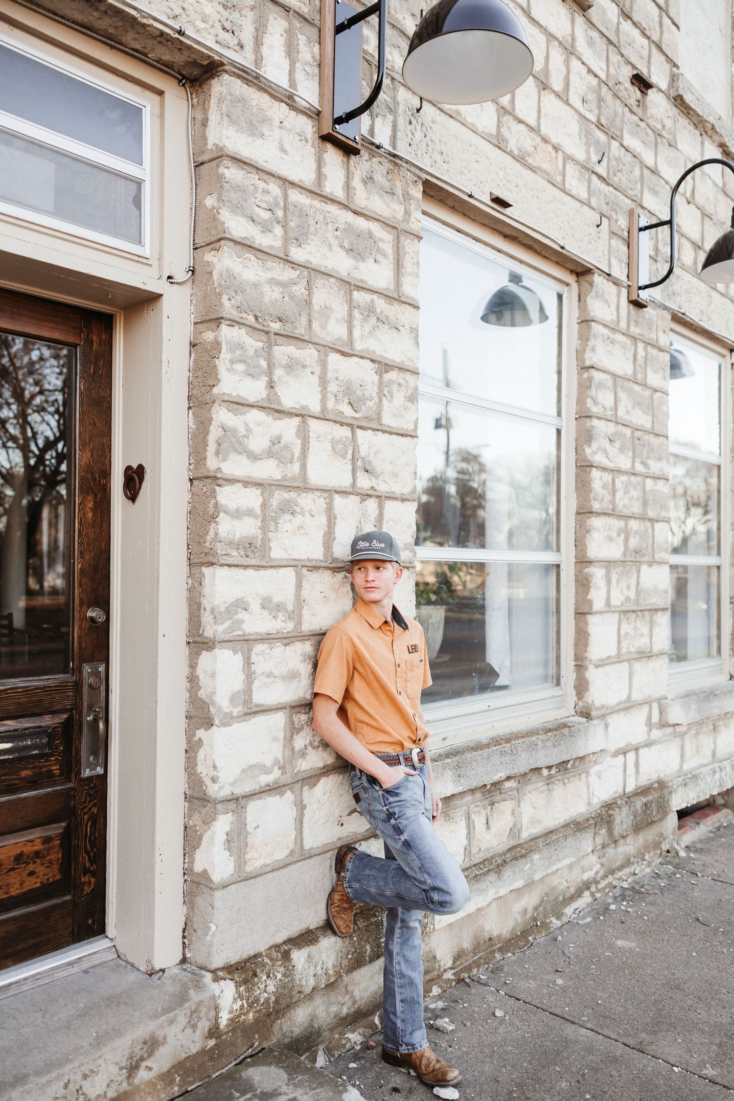 Man in orange shirt and cap leans against rustic stone building facade on urban sidewalk.