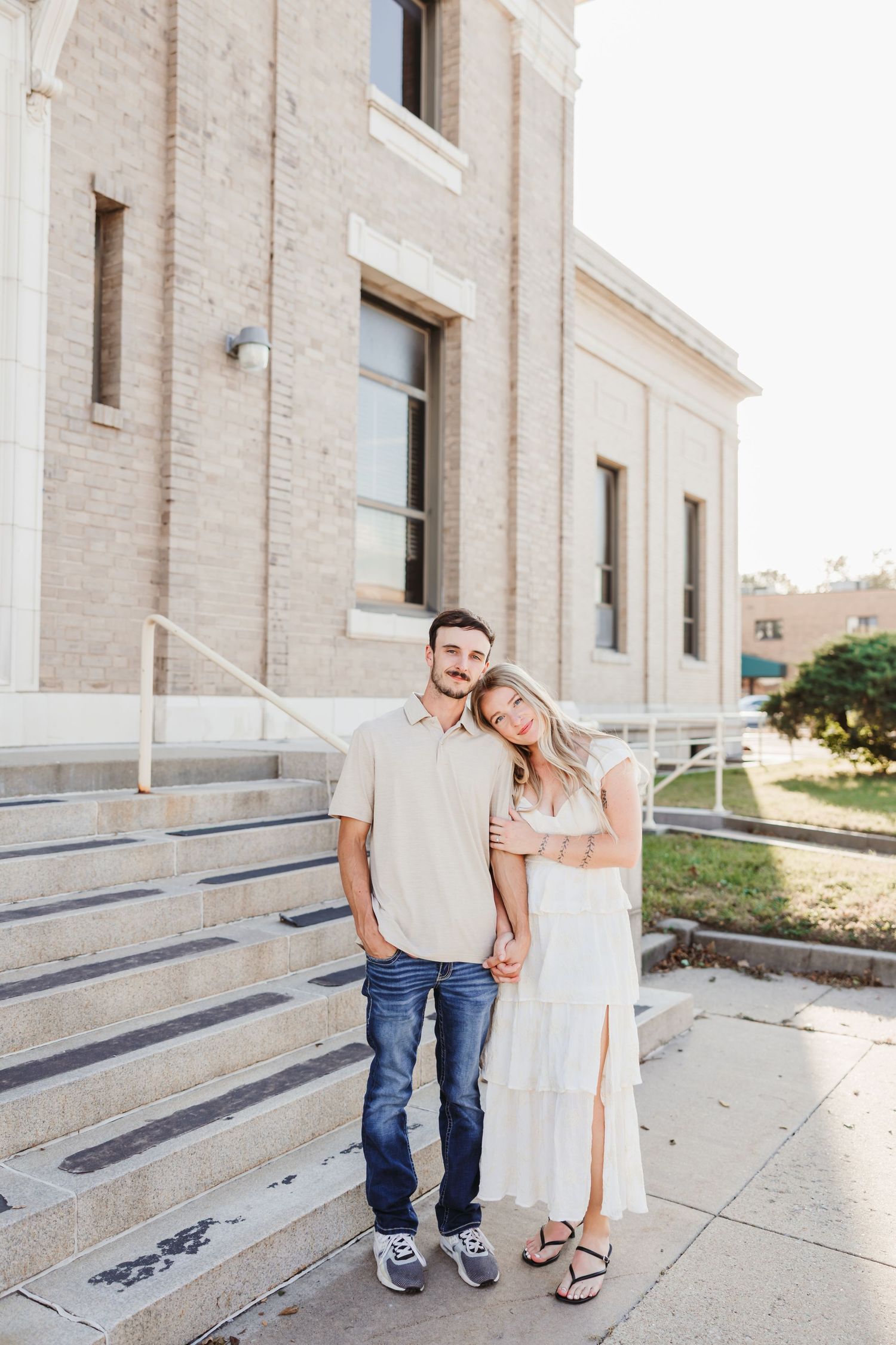 Couple posing on concrete steps outside old historic building in casual attire.