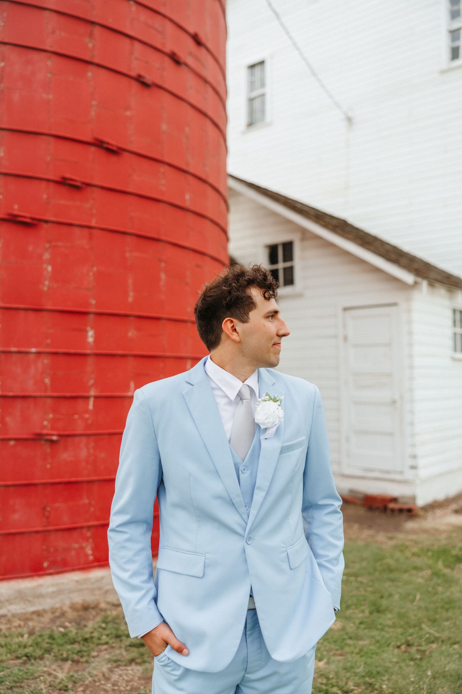 Groom in a light blue suit with boutonniere stands confidently in front of a red silo.