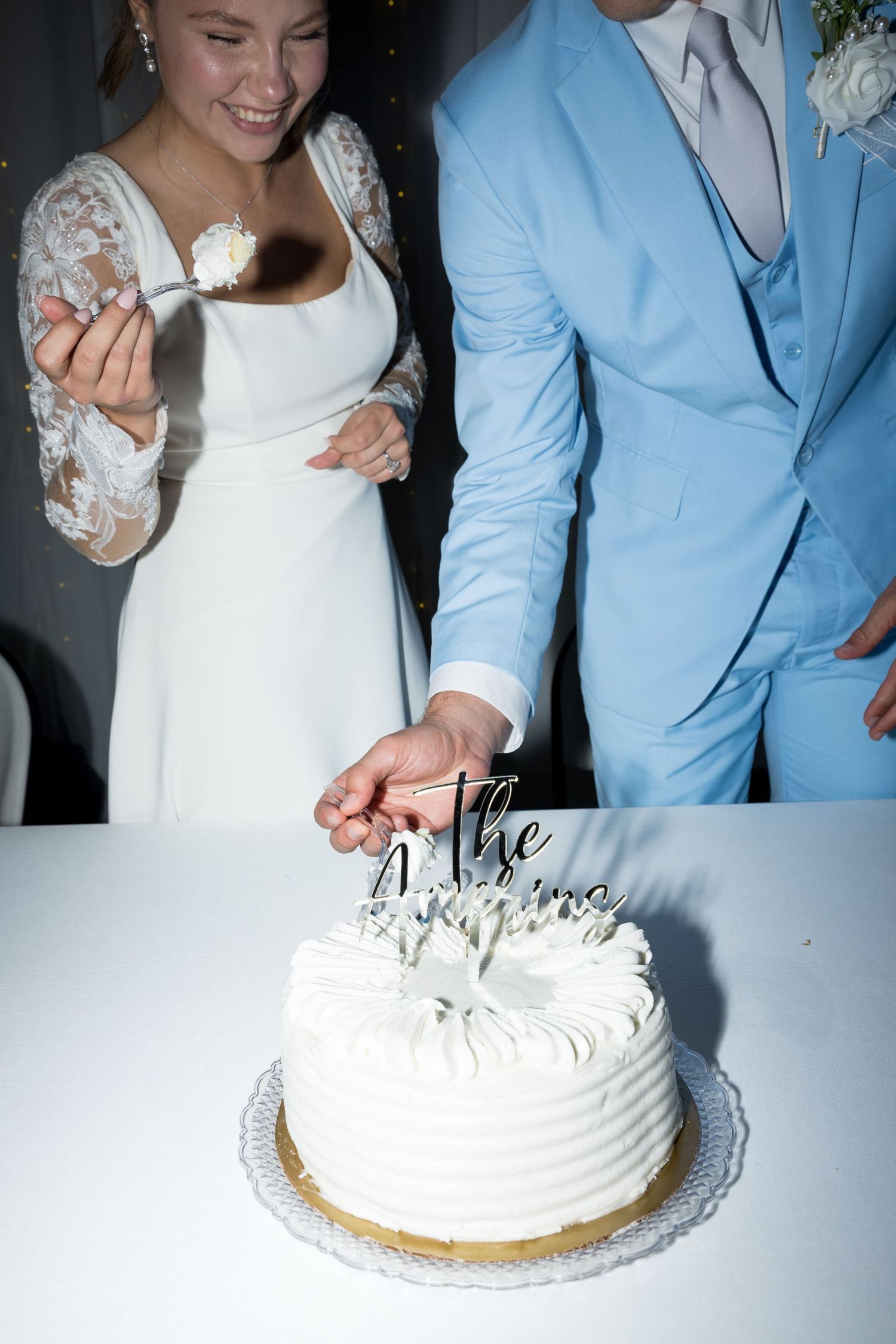 Bride in white lace dress and groom in light blue suit cutting white wedding cake together.