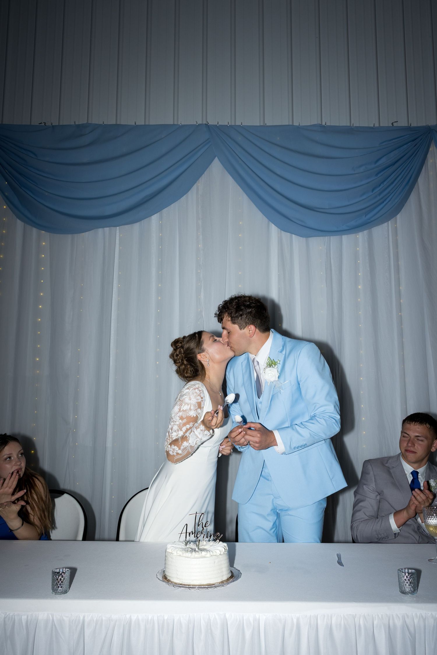 Newlyweds share a romantic kiss behind their wedding cake at reception with blue draped backdrop.