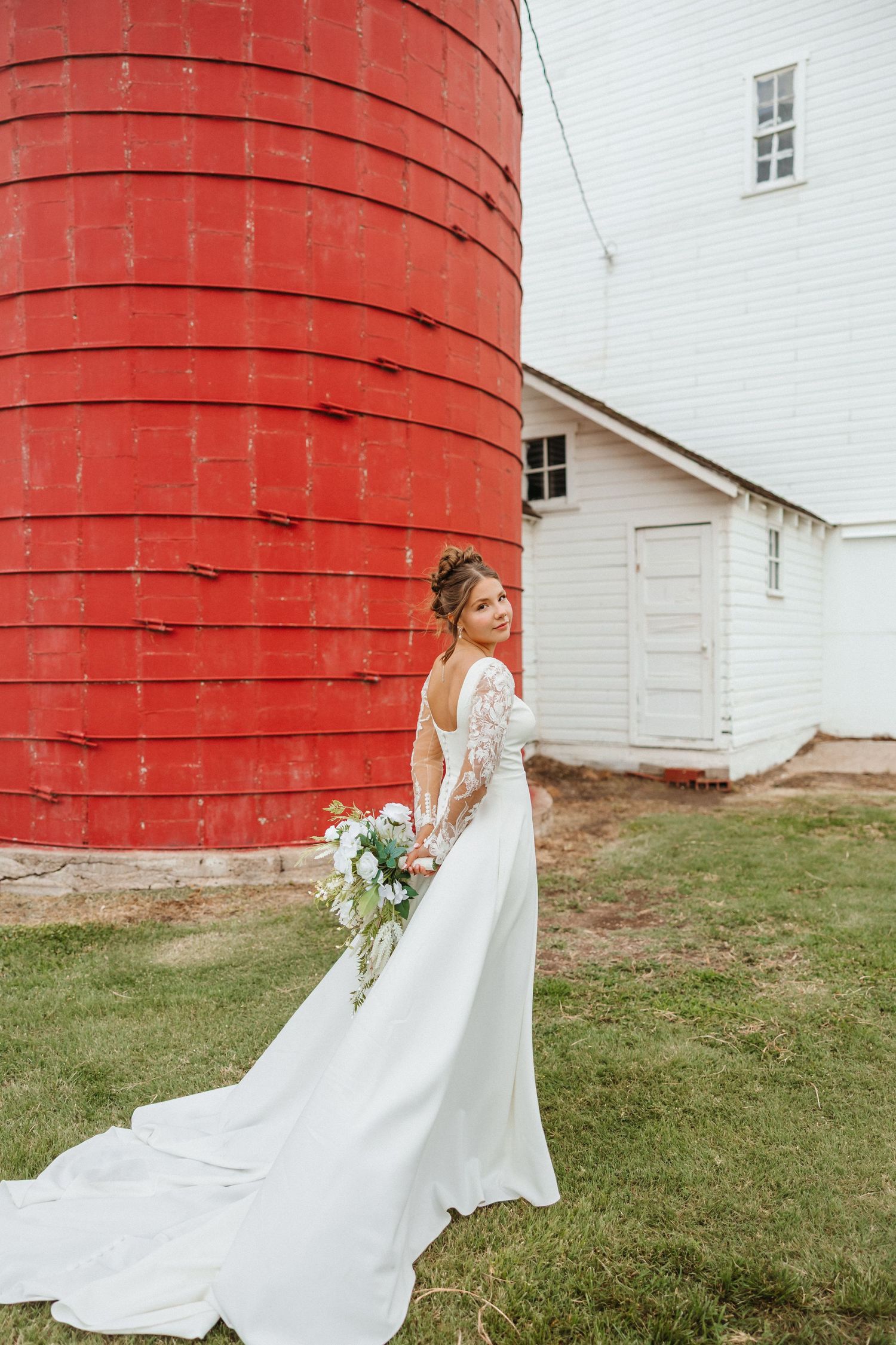 Bride in a long-sleeved lace wedding gown holds a bouquet while posing beside a red silo.