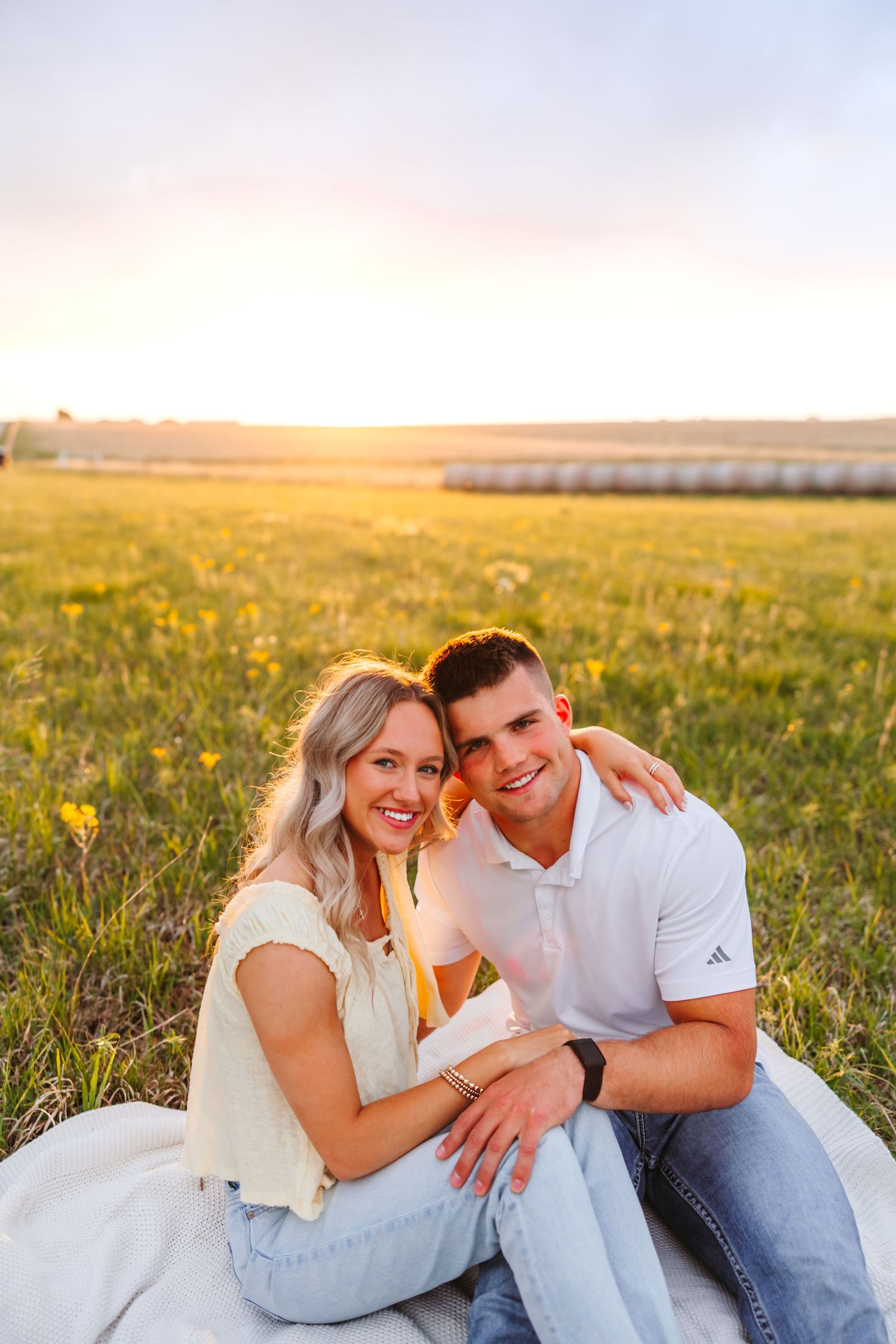 A smiling couple sits together on a blanket in a sunny field at golden hour, with hay bales in the background.