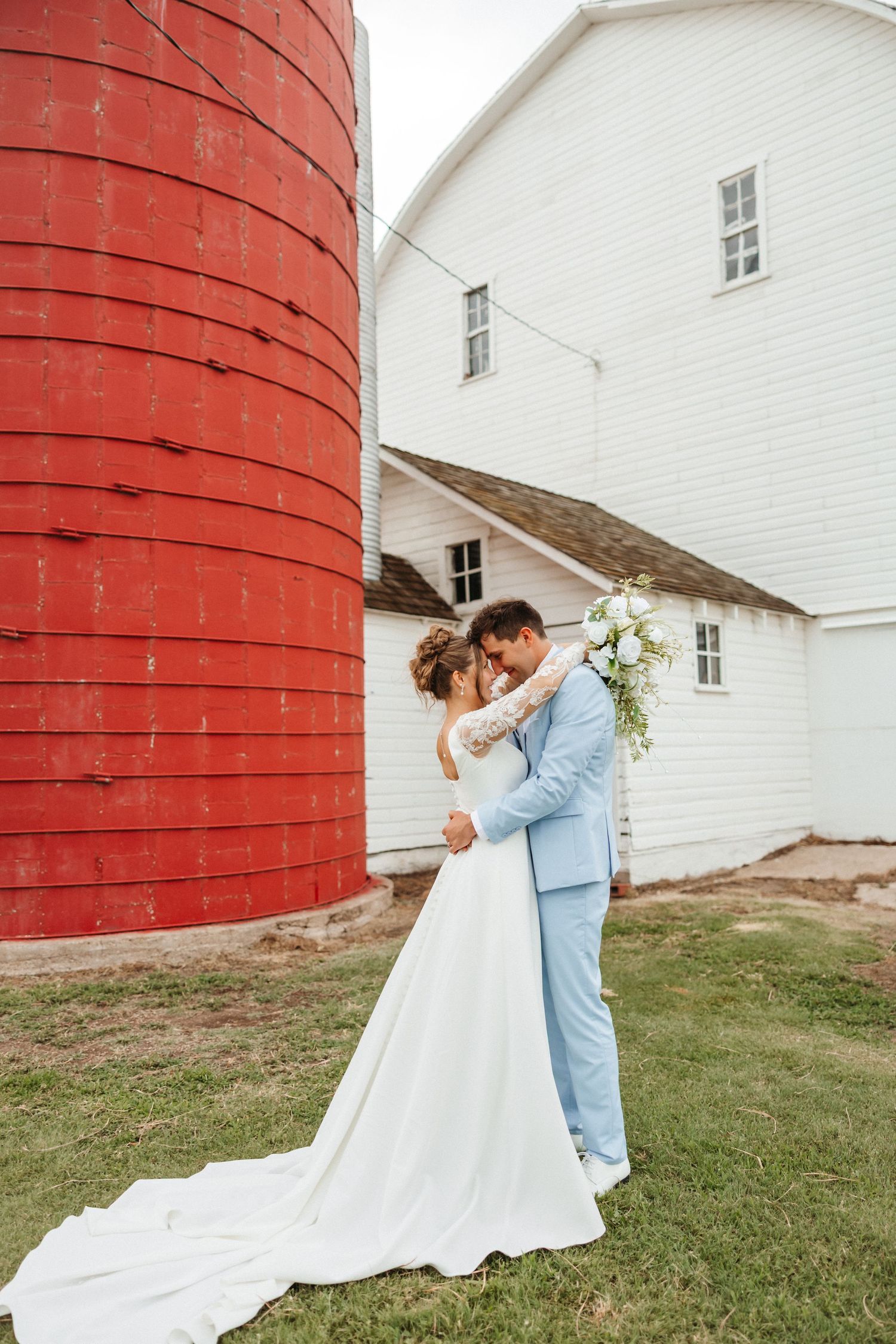 Bride and groom embrace near a red silo and white barn on a farm wedding day.