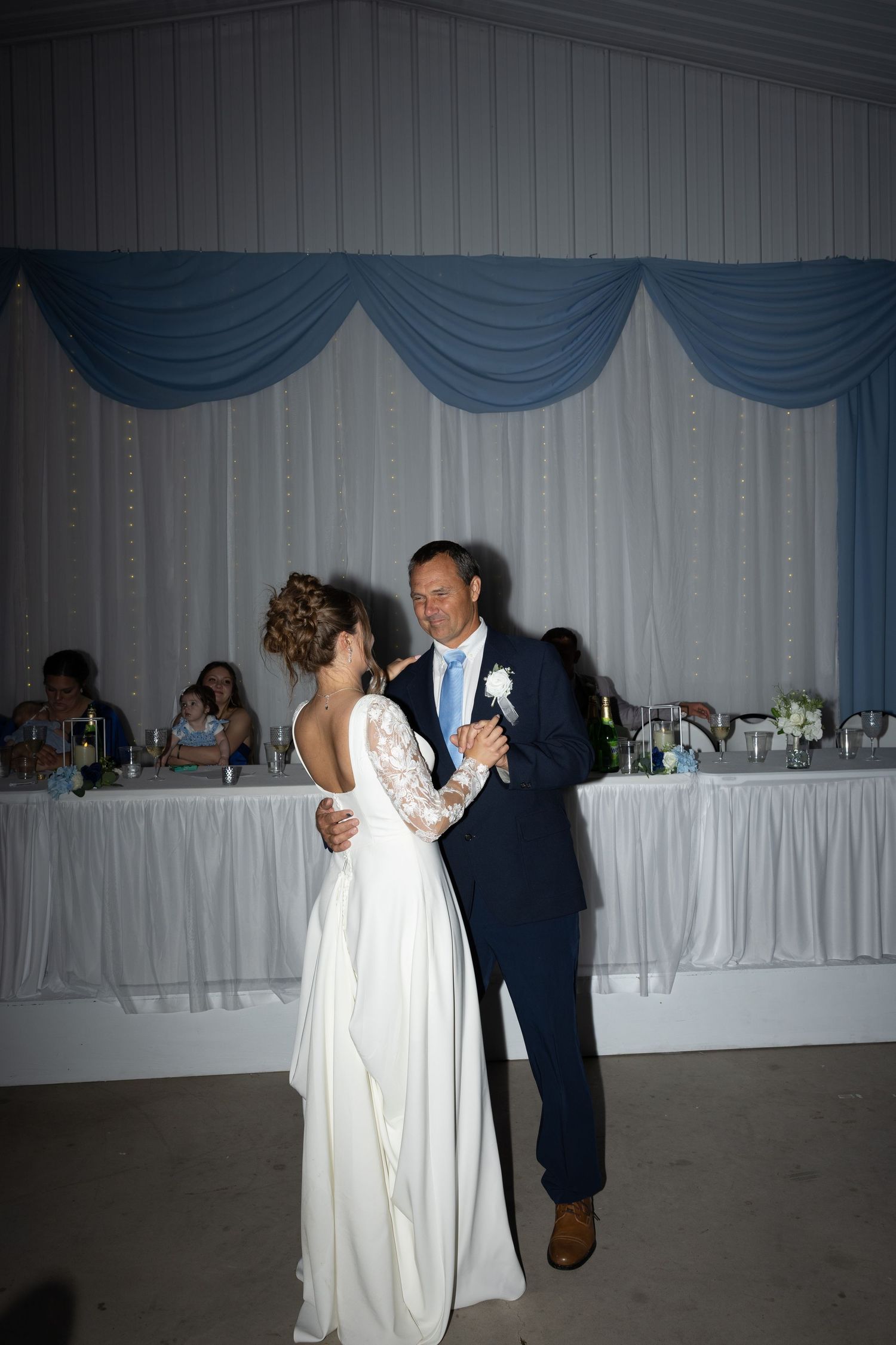 Bride and groom share their first dance at wedding reception with blue draped backdrop and guests seated at head table.