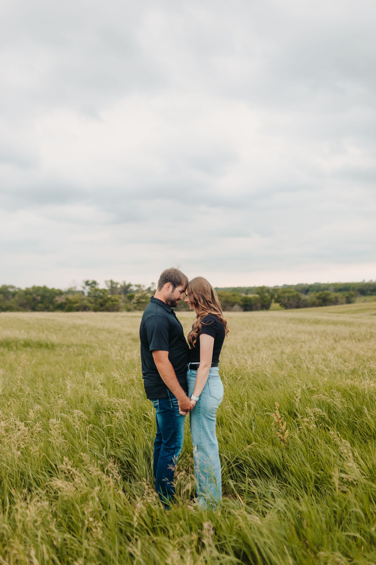 A couple holds hands and touches foreheads while standing in a lush green field under a cloudy sky.